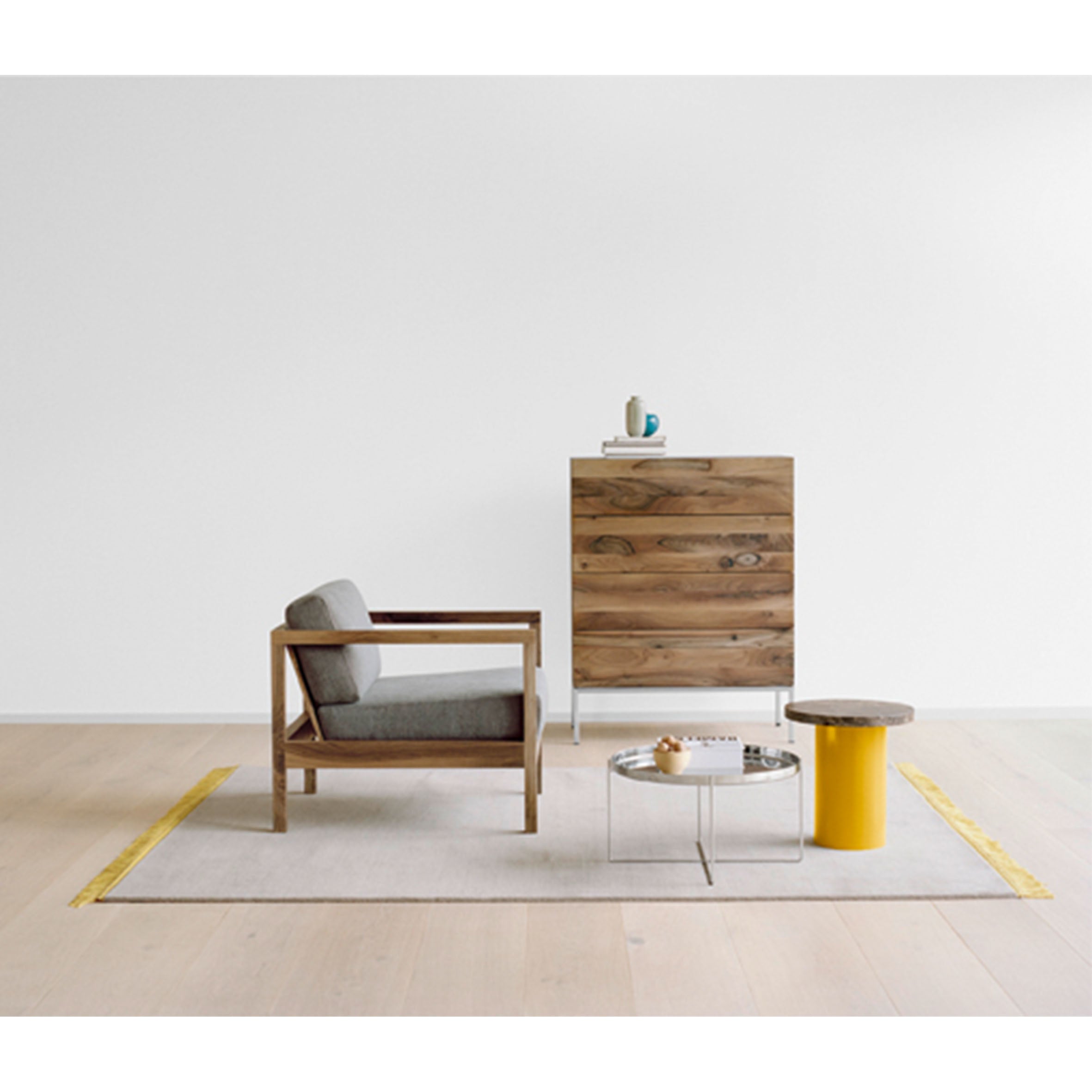 A minimalist room features a wooden armchair, gray cushions, and the E15 EC01 BYRON by a sleek yellow side table. A modern metal coffee table with books and a wooden cabinet complements the white wall. The light wood floor is adorned with a beige rug.