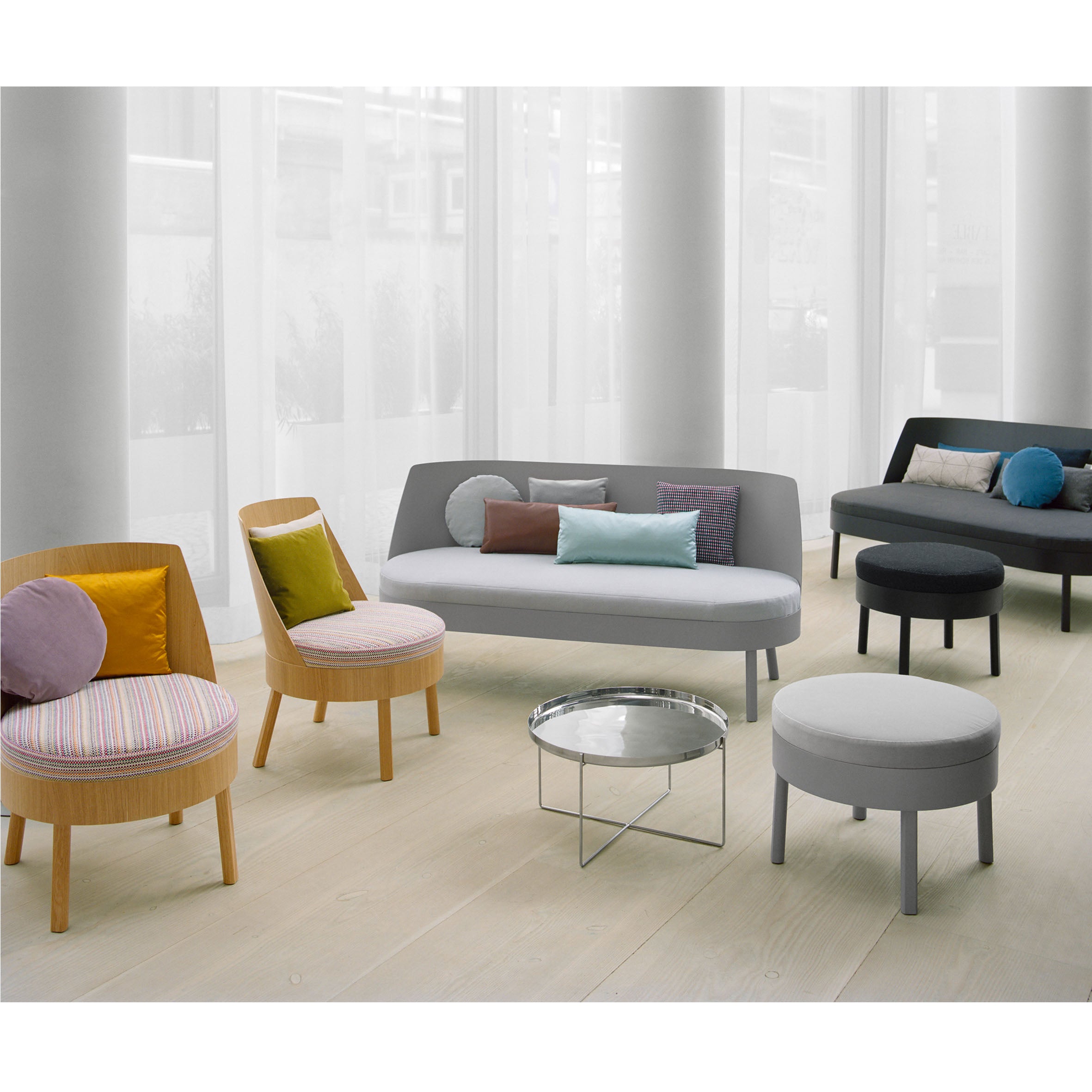 A modern room with oak veneer plywood flooring includes three small couches with colorful pillows, two light wooden chairs with patterned cushions, a round glass-top table, and a gray ottoman. E15s sleek EC02 BESSY Lounge Chair complements the tall white curtains in the background.