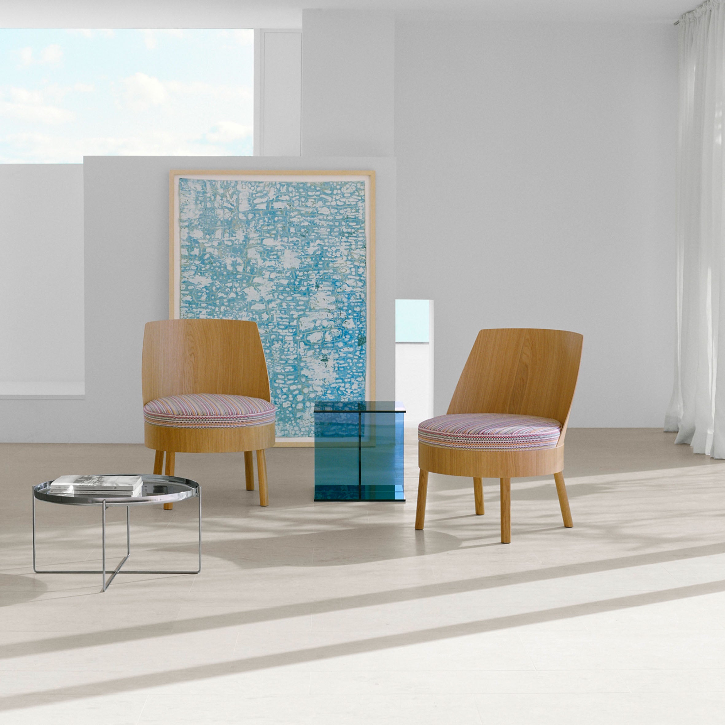 In a minimalist room, two E15 EC02 BESSY Lounge Chairs with striped cushions face a low round table to the left, while a blue glass cube sits centrally. A large abstract artwork leans against the wall near a window, and soft curtains on the right enhance the serene ambiance.