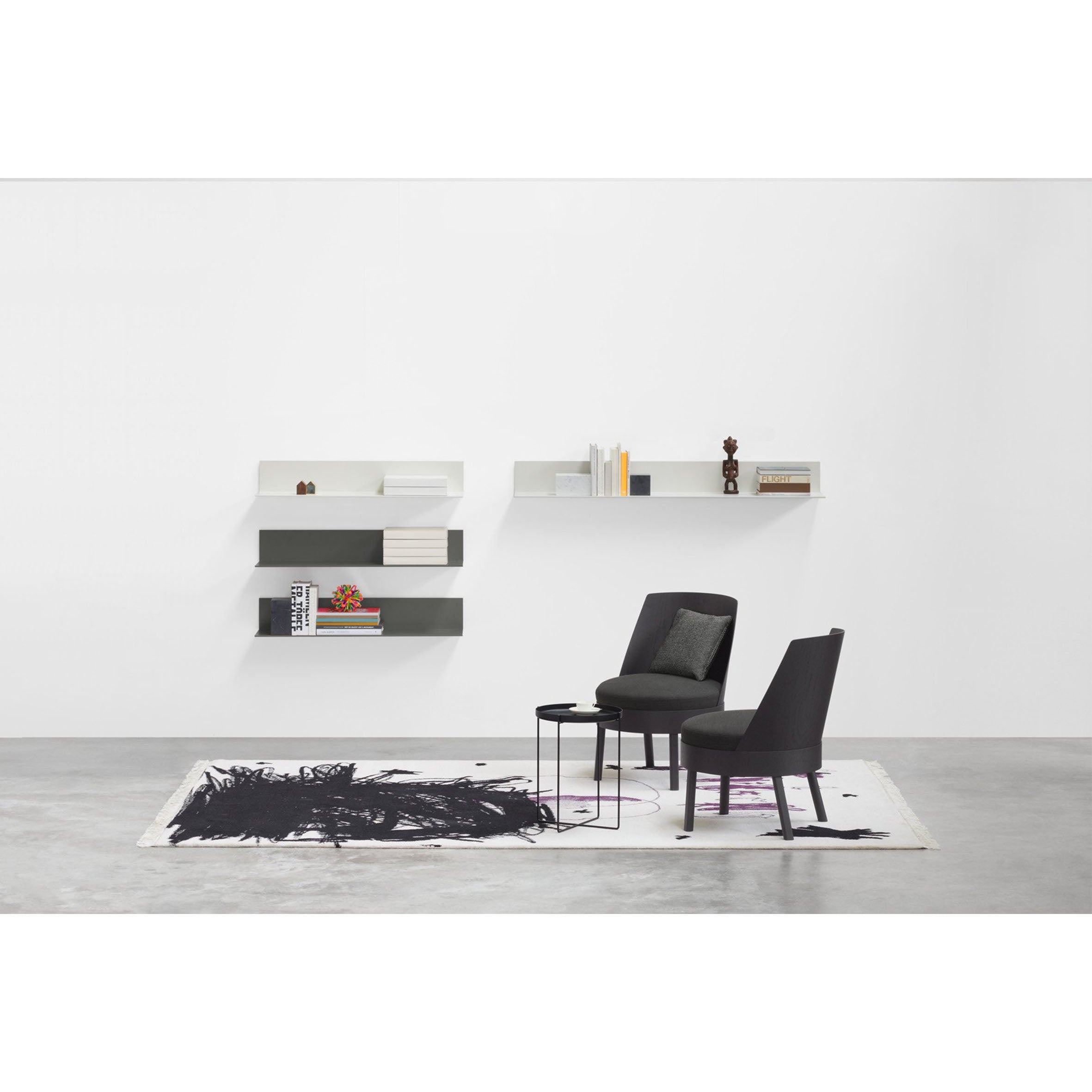 A minimalist interior features two EC02 BESSY Lounge Chairs by E15 and a round side table on a black and white abstract rug. Oak veneer plywood shelves display books and decor against the white wall, with a concrete floor completing the look.