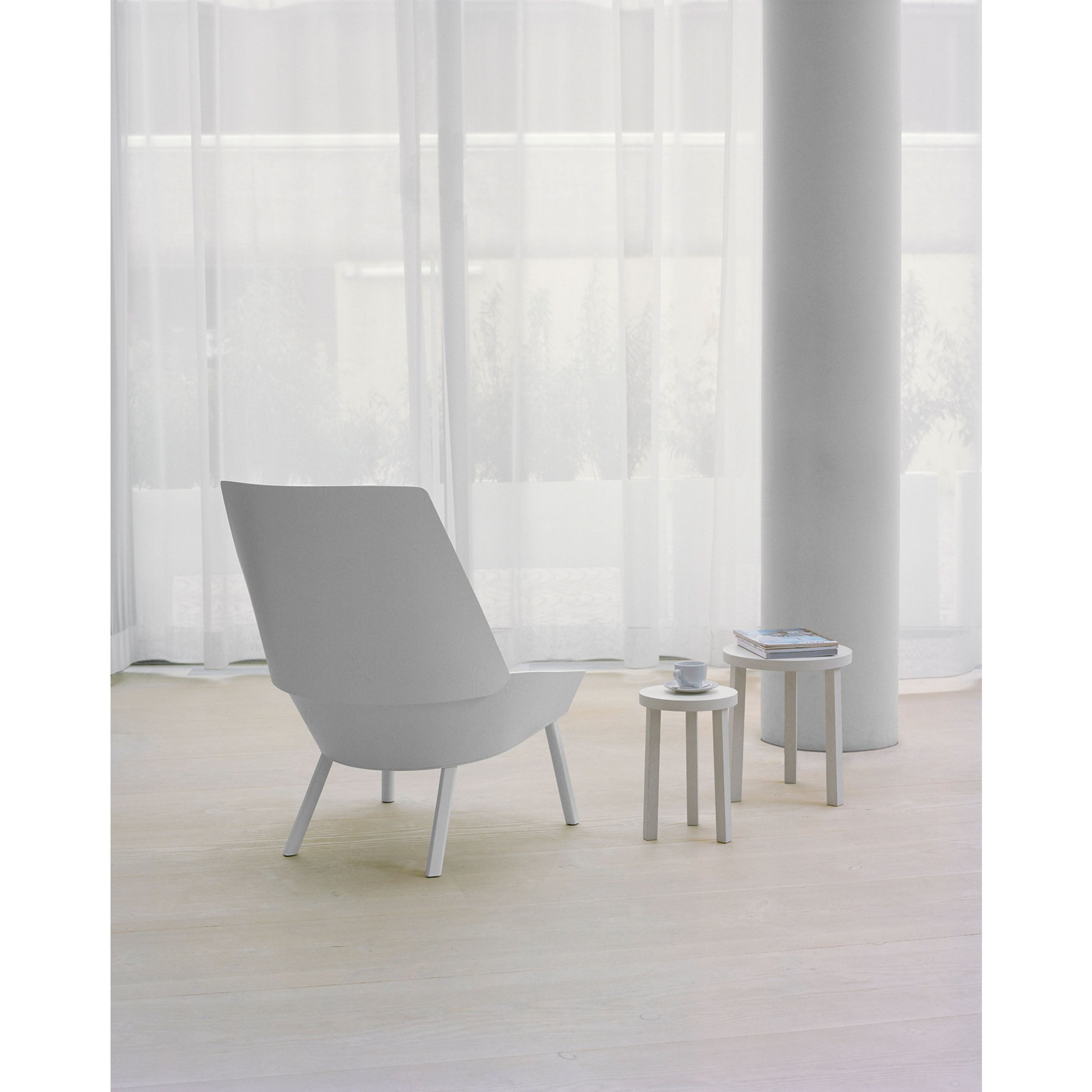A minimalistic room showcases E15s EC03 EUGENE modern gray lounge chair, alongside two small white tables on a light wooden floor. One oak veneer-finished table holds books, the other a cup and saucer, with large sheer curtains allowing soft natural light in.