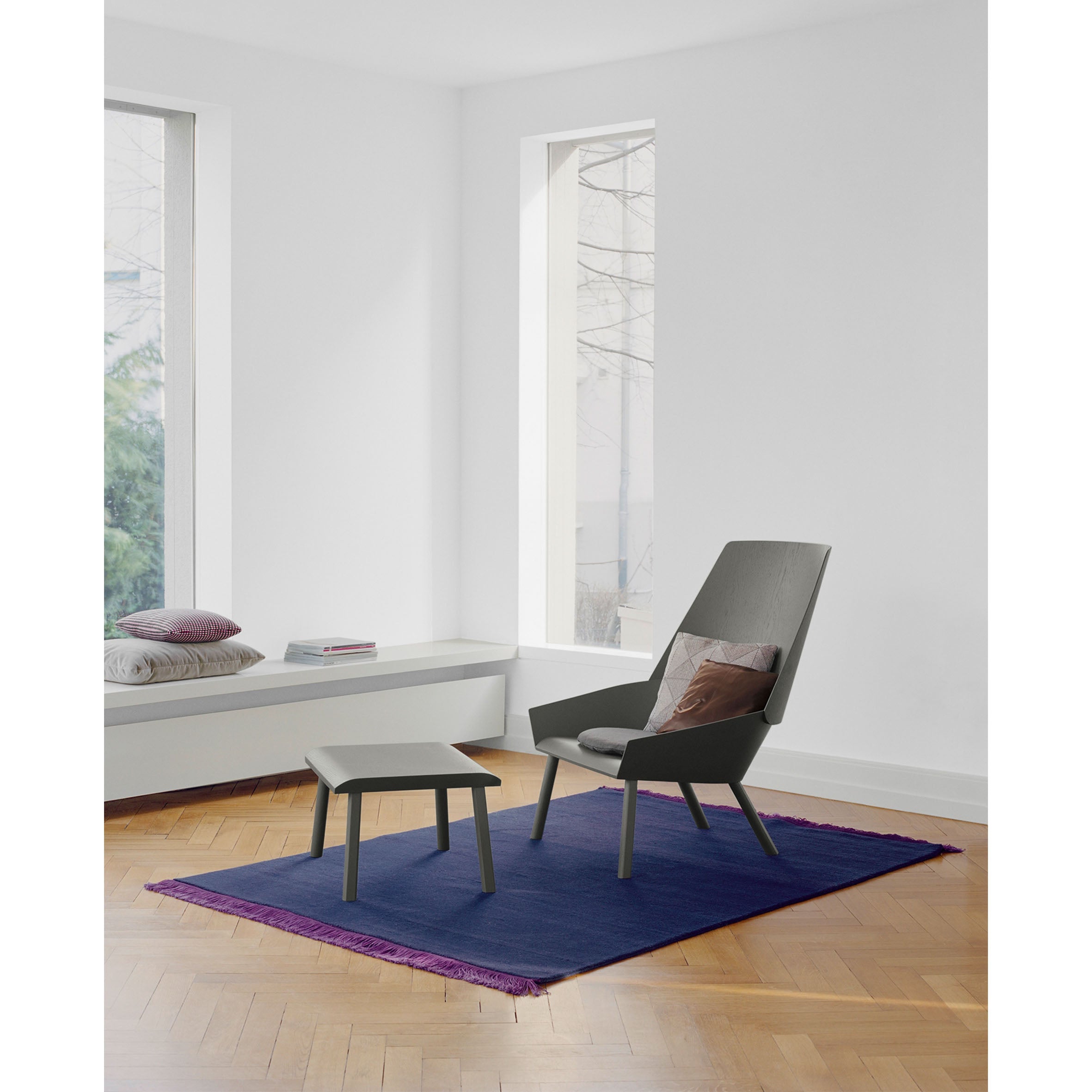 A minimalist room features the EC03 EUGENE gray lounge chair and matching ottoman from E15 on a fringed purple rug. Natural light streams through large windows, highlighting an oak veneer wooden floor and a bench with cushions and a book.