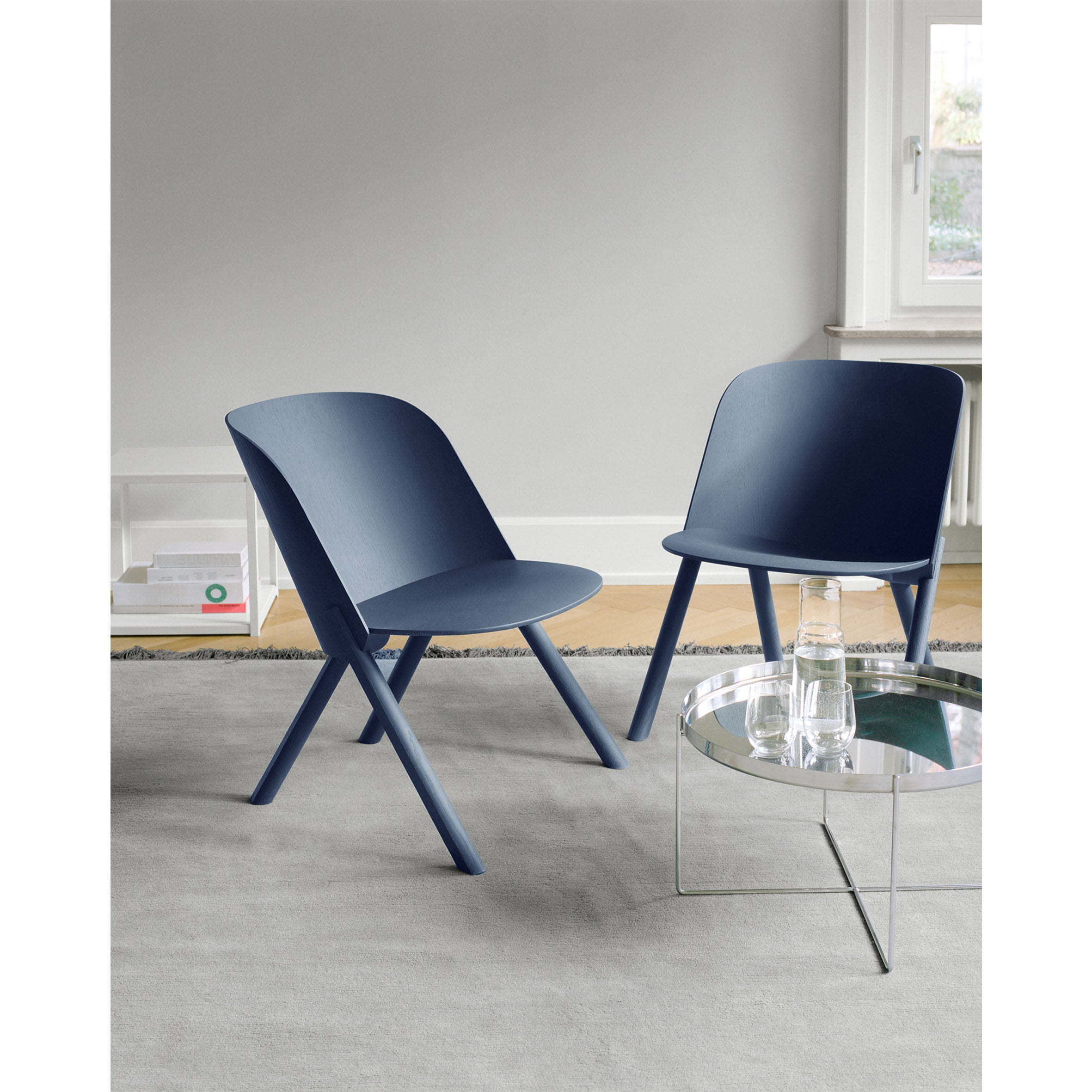 Two modern blue EC05 THAT lounge chairs from E15 with angled legs rest on a light gray carpet. Nearby, a round glass-top coffee table holds a water carafe and glass. The room is softly illuminated by natural light from the window.