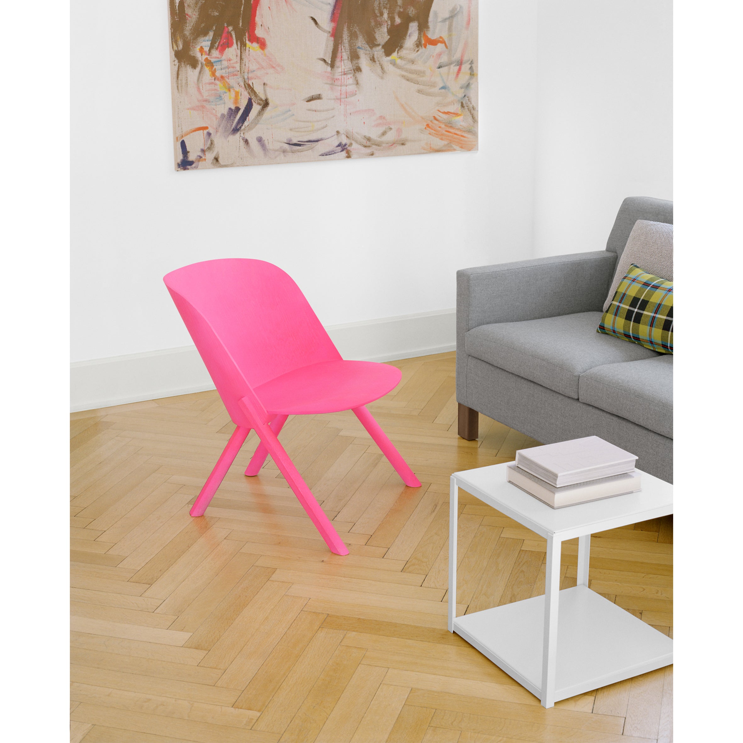 The E15 EC05 THAT pink upholstered seat is on a wooden herringbone floor next to a gray sofa with a plaid cushion. Nearby, theres a white side table with stacked books and an abstract painting on the white wall above.