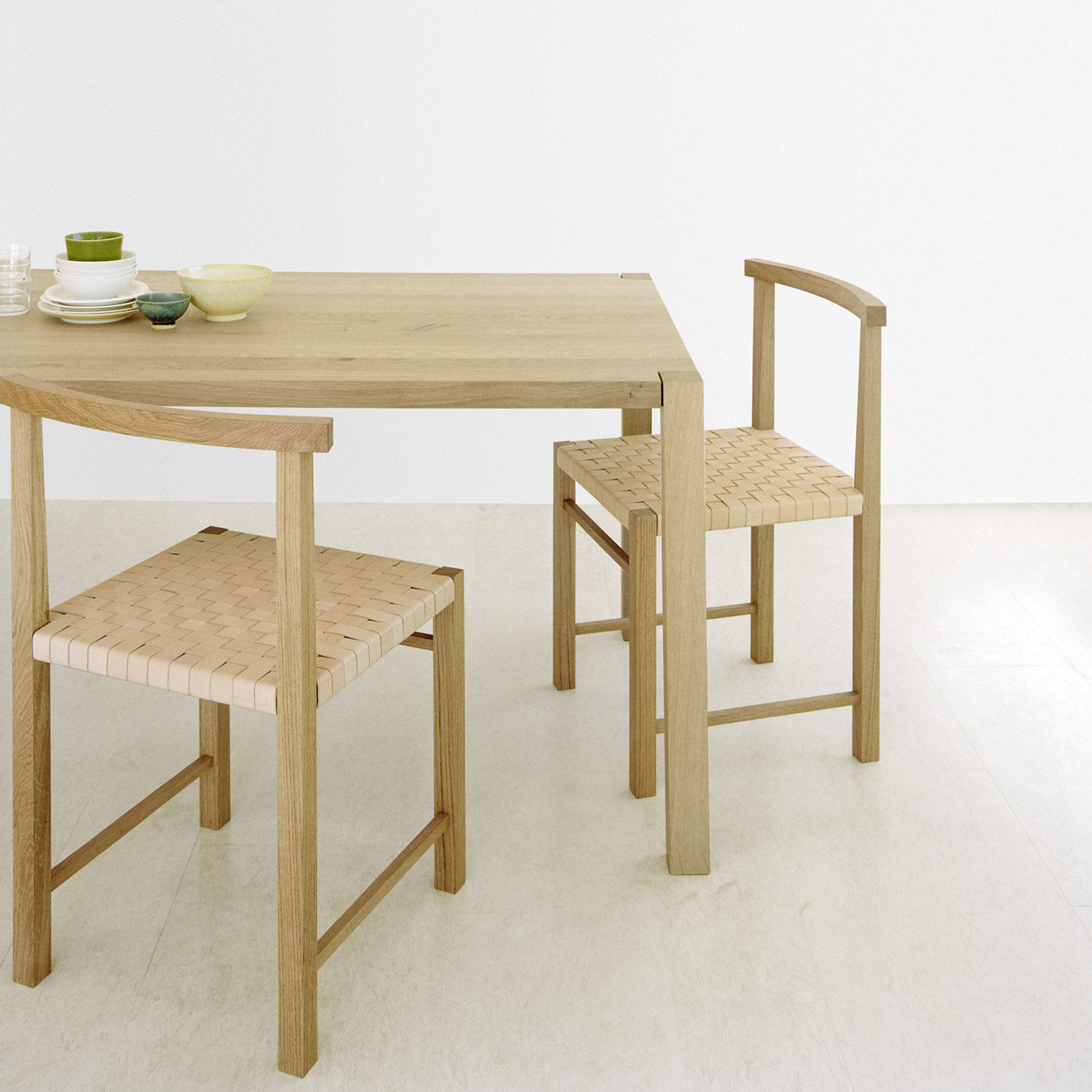 The FK02 KARNAK dining set by E15 features a light wood table with Ferdinand Kramer chairs. A small stack of plates and bowls adorns the table in a bright, neutral-toned room, highlighting the elegance of waxed oak in a simple, modern design.