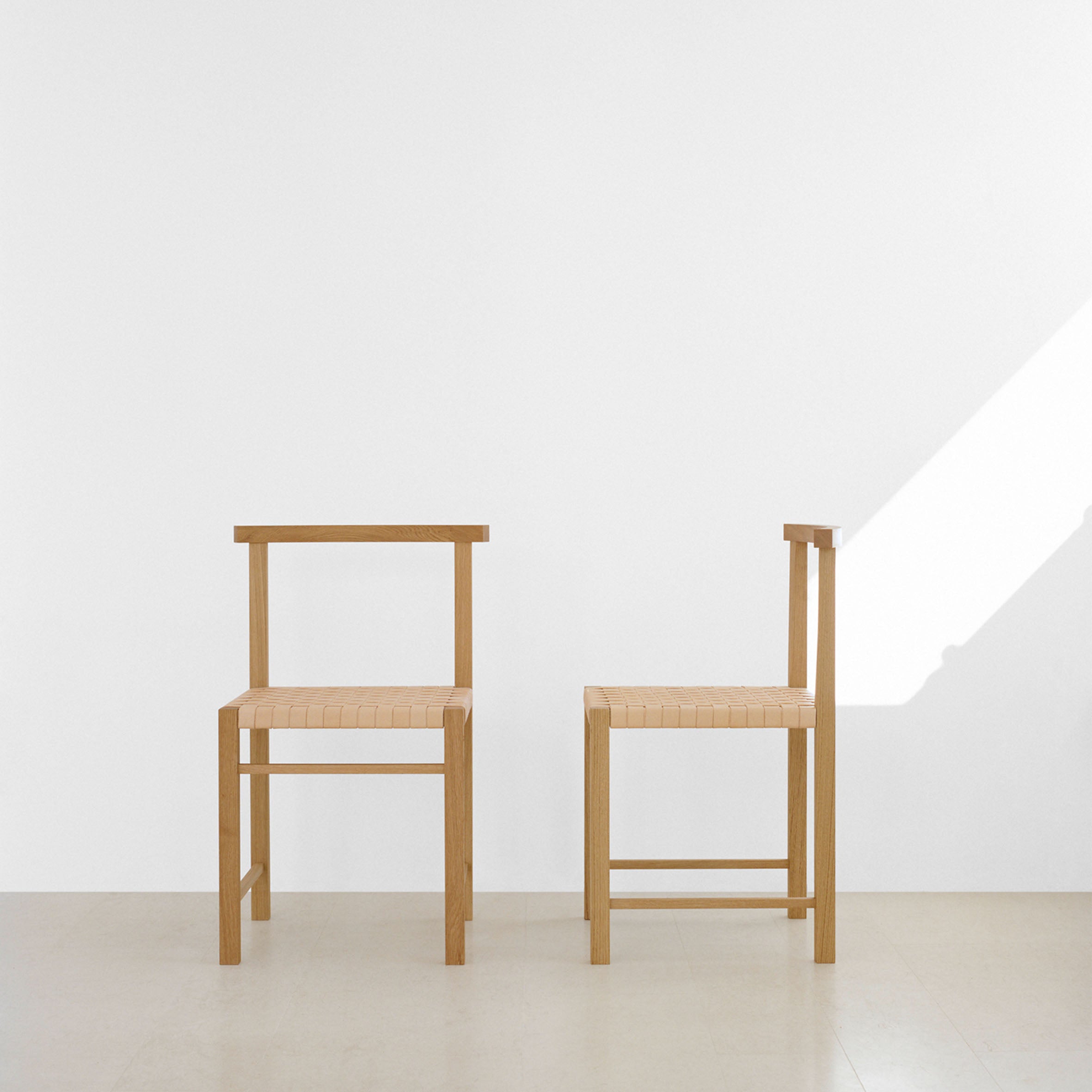Two FK02 KARNAK chairs by E15, in a minimalist design reminiscent of Ferdinand Kramer, sit against a white wall. Natural light casts shadows enhancing the settings simplicity. The flooring is neutral beige, contributing to the modern aesthetic.