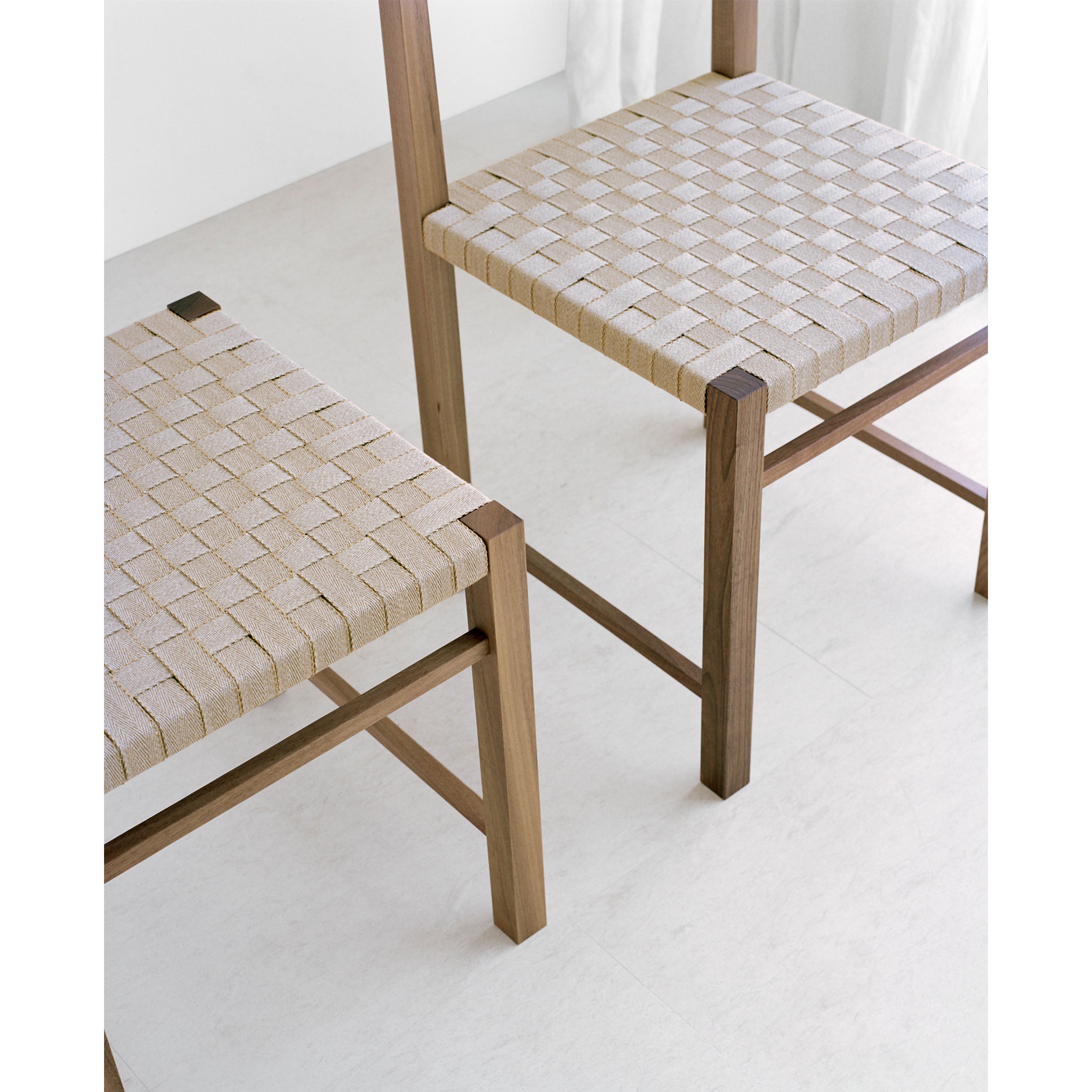 Two waxed oak FK02 KARNAK chairs by E15 feature beige woven seats. Set on a light floor with a curtain in the background, the design is minimalist and modern.