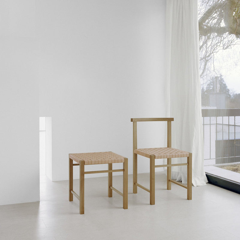 Two FK03 ASWAN chairs by E15, featuring minimalist wooden design and handwoven seats, are set against white walls and flooring. A large window with sheer curtains allows natural light to flood in, highlighting the waxed solid oak finish of the furniture against a backdrop of trees.
