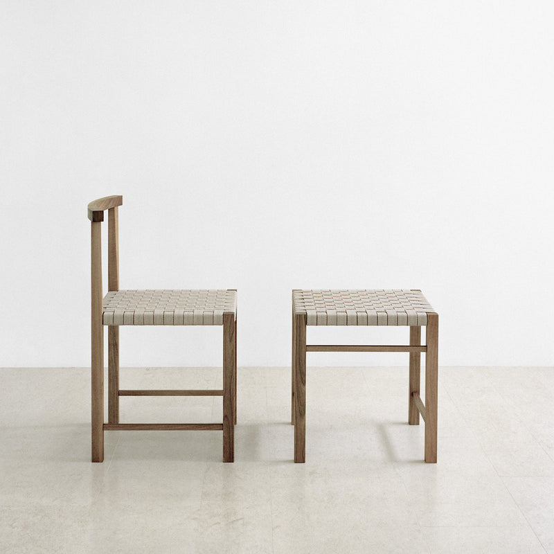 Two minimalist chairs with waxed solid oak frames and handwoven seats sit on a light-colored floor against a plain white background—one with a backrest and the other, E15’s FK03 ASWAN stool, highlighting its backless design.