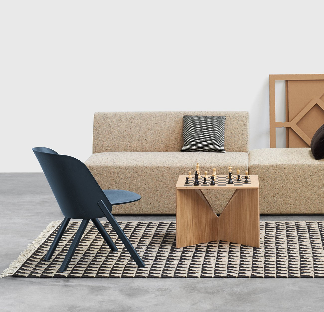 In the modern living room, a beige sofa pairs with a dark gray chair, while an FK04 CALVERT Chess Board by E15 graces the wooden coffee table. A geometric-patterned rug adorns the floor, complemented by a decorative panel leaning against the wall and an artfully placed gray pillow on the sofa.