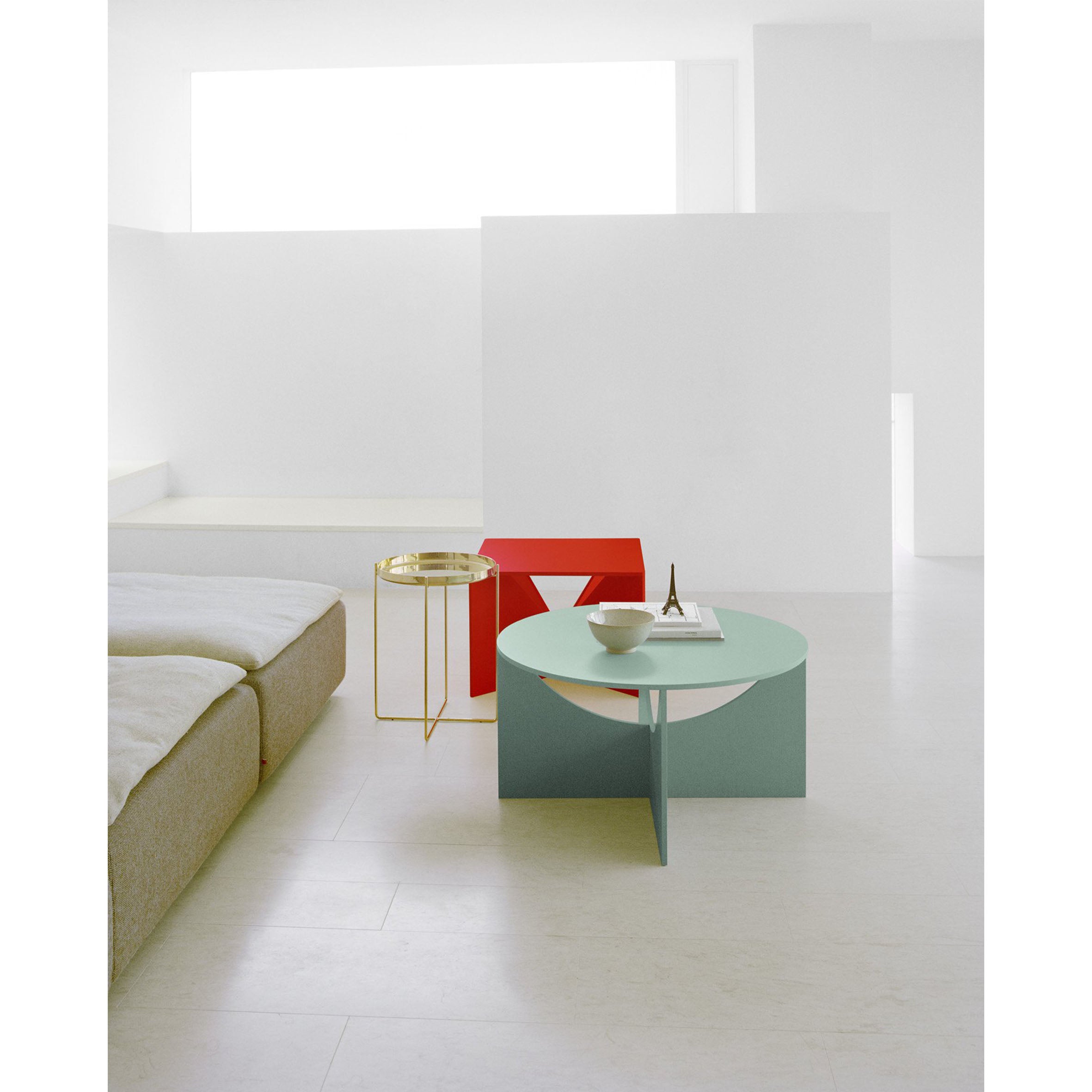 A minimalist room features a beige sofa and blue circular table with a small vase, complemented by the E15 FK05 CHARLOTTE coffee table designed by Ferdinand Kramer. A gold round side table adds elegance, while white walls and soft light from a large window create an inviting ambiance.