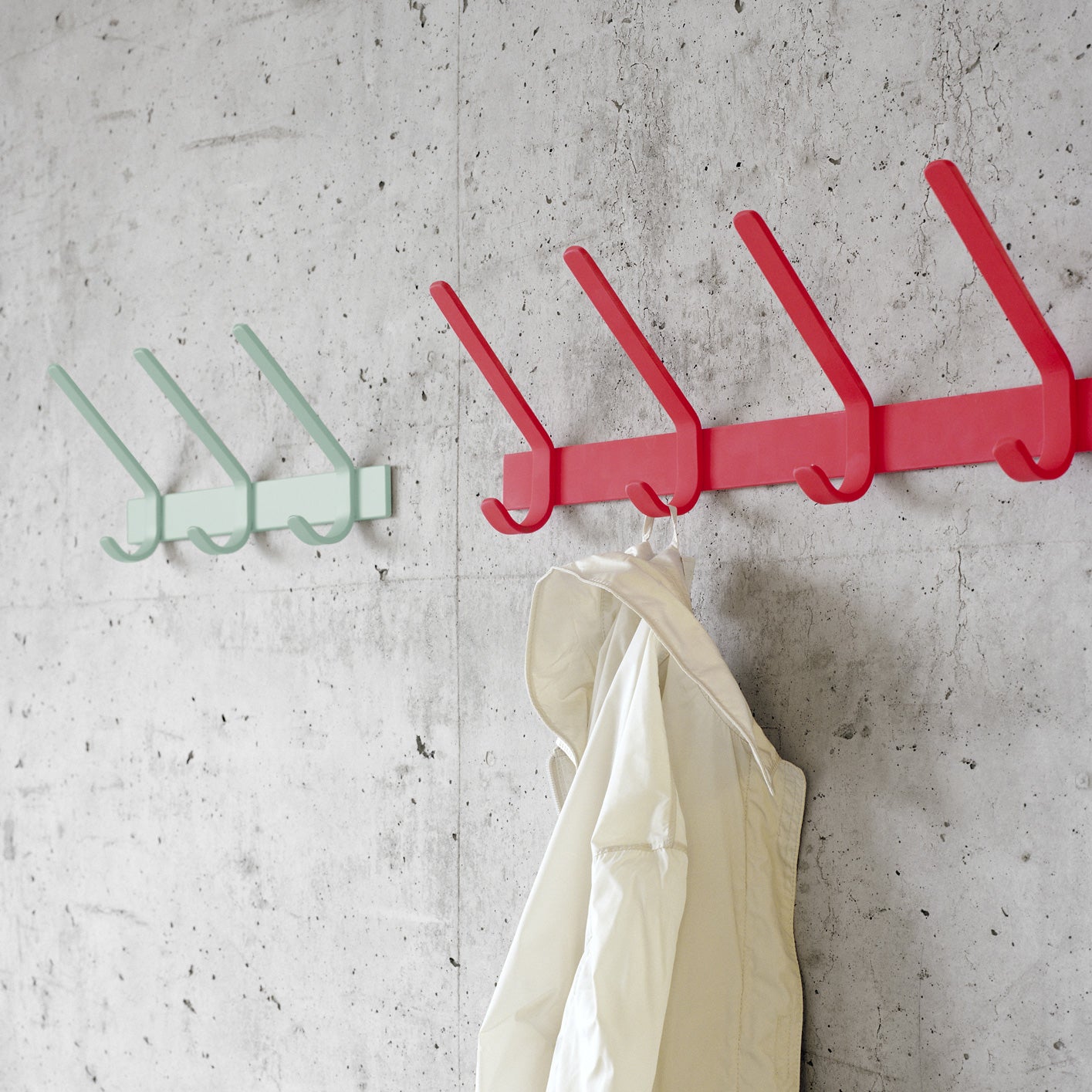 Two FK08 UNI coat racks by E15, made of powder-coated steel, are mounted on a concrete wall: one red with five hangers holding a white jacket and the other mint green with three empty hangers. Their sleek, minimalist design contrasts with the industrial backdrop, evoking Ferdinand Kramers style.