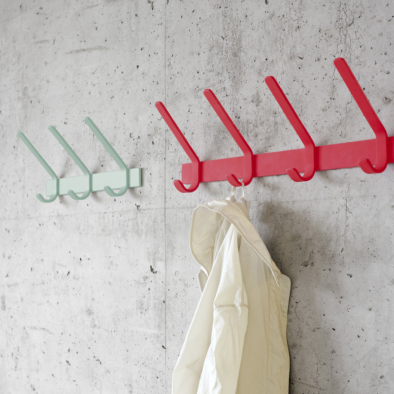 Two FK08 UNI coat racks by E15, made of powder-coated steel, are mounted on a concrete wall: one red with five hangers holding a white jacket and the other mint green with three empty hangers. Their sleek, minimalist design contrasts with the industrial backdrop, evoking Ferdinand Kramers style.