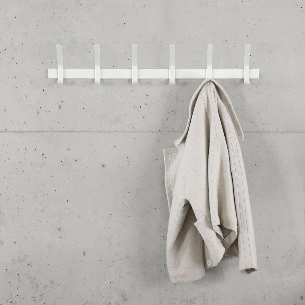 The FK08 UNI coat rack by E15, crafted in minimalist powder-coated steel reminiscent of Ferdinand Kramers designs, is mounted on a gray concrete wall with one empty hook and another holding a light beige coat.