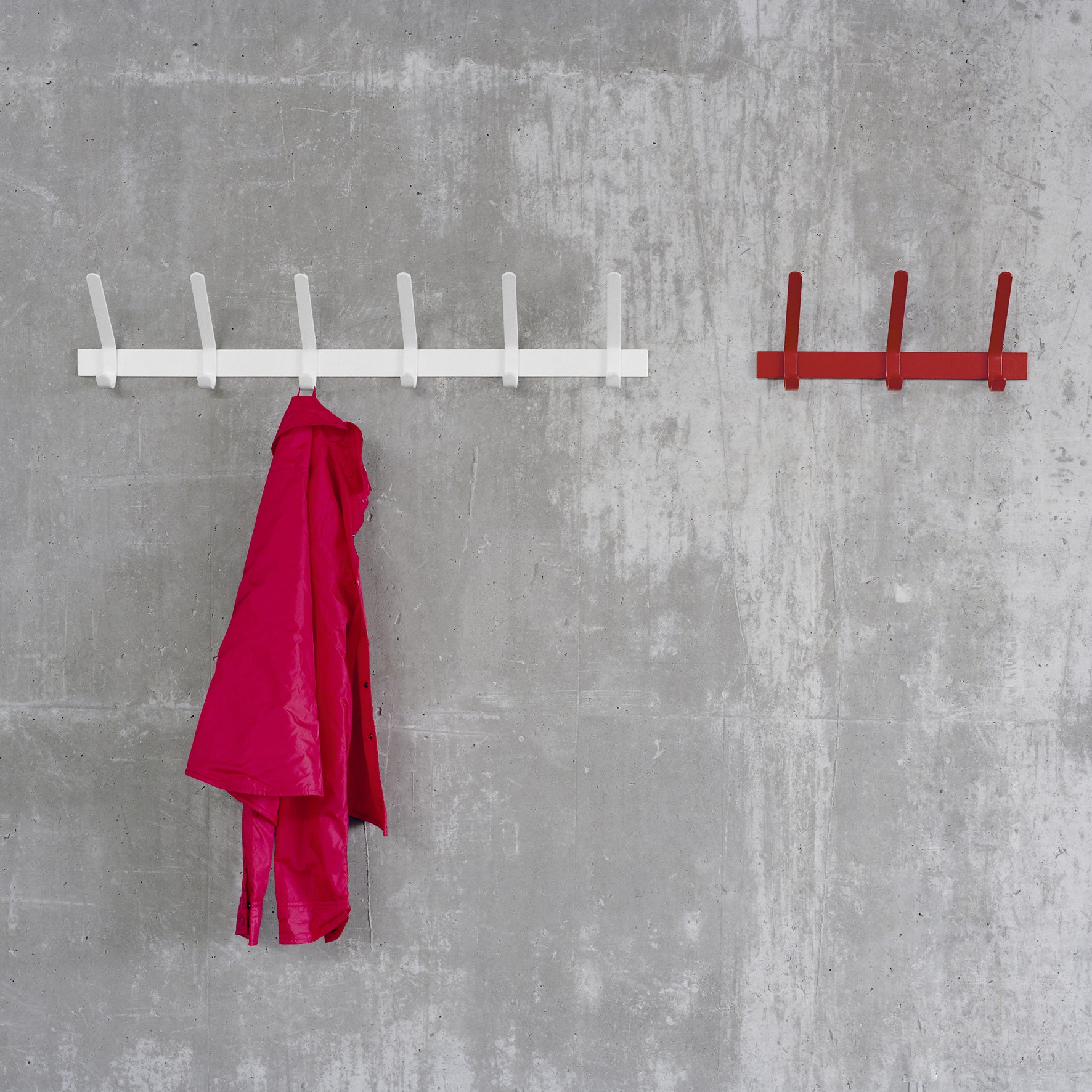 A textured concrete wall showcases two mounted coat racks. On the left, E15s FK08 UNI by Ferdinand Kramer is white with a bright pink coat hanging from one hook. The smaller red rack on the right, made of powder-coated steel, is bare.