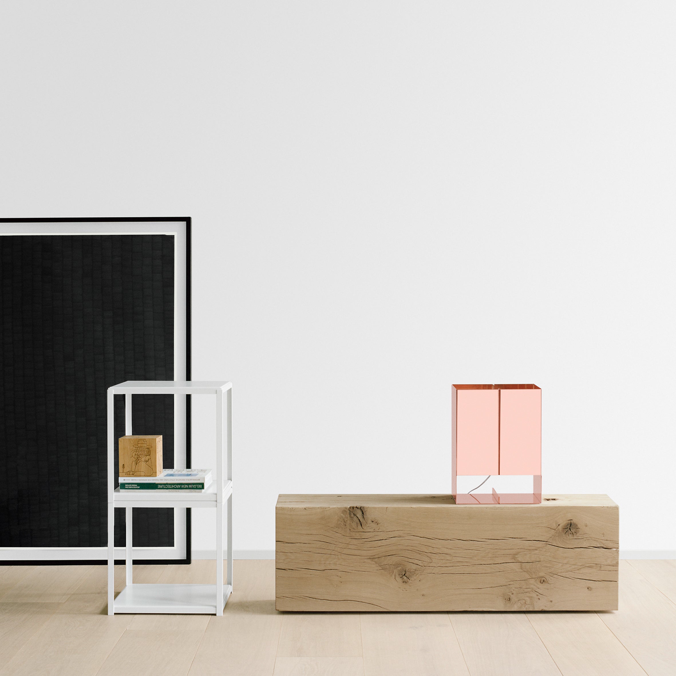 A minimalist room features a pink glass lamp on a light wood block on the right. To the left, E15s FK12 FORTYFORTY, in powder-coated steel, functions as a small white shelf holding books and a framed picture. A large black-framed artwork leans against the white walls over the light wood floor.
