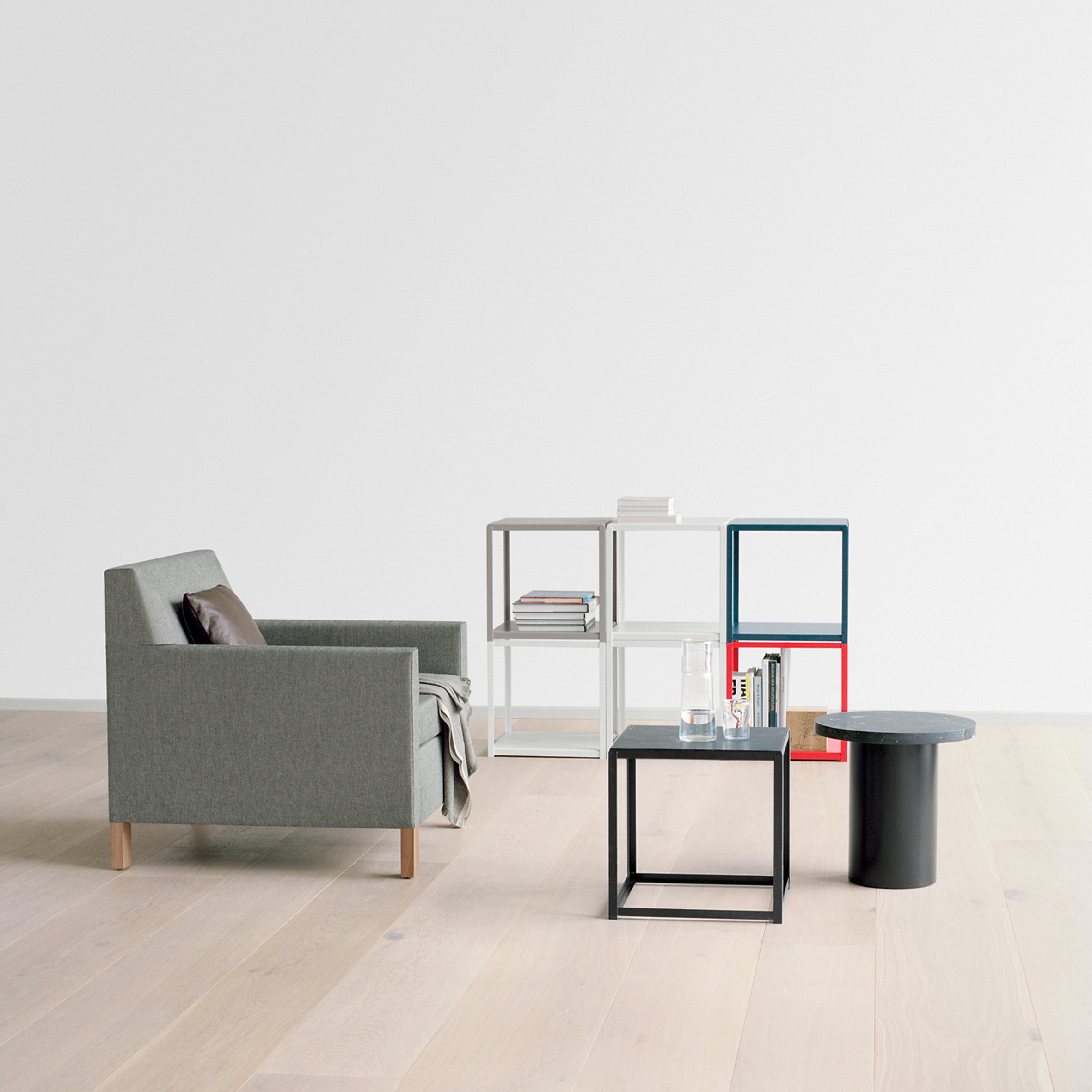 Minimalist living room featuring a gray armchair and E15s FK12 FORTYFORTY geometric side tables in powder-coated steel on light wood flooring. The tables hold books, a glass, and a small container, set against a blank white wall that emphasizes the clean design.