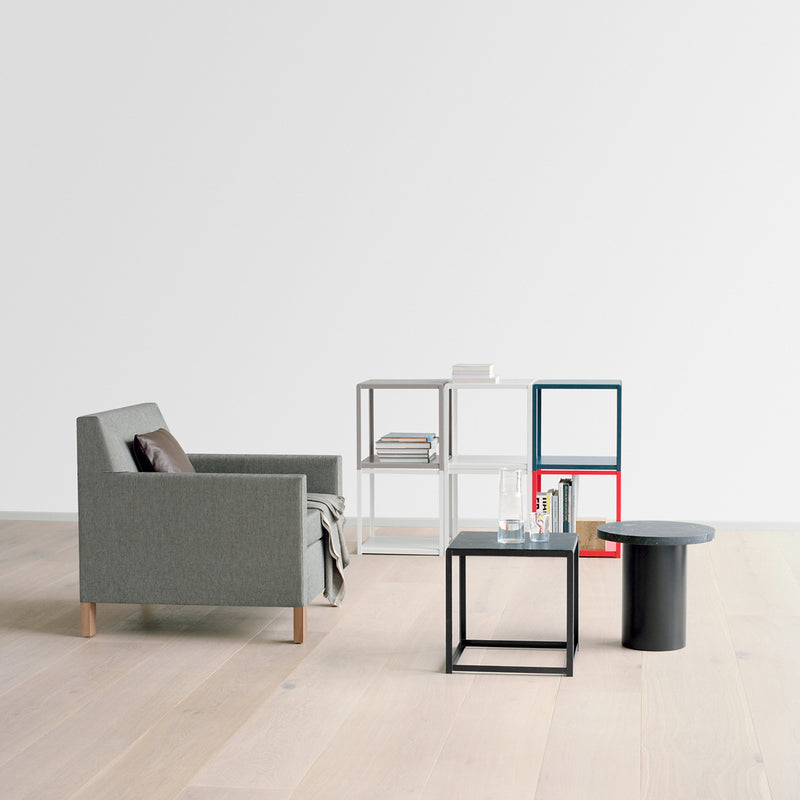 Minimalist living room featuring a gray armchair and E15s FK12 FORTYFORTY geometric side tables in powder-coated steel on light wood flooring. The tables hold books, a glass, and a small container, set against a blank white wall that emphasizes the clean design.