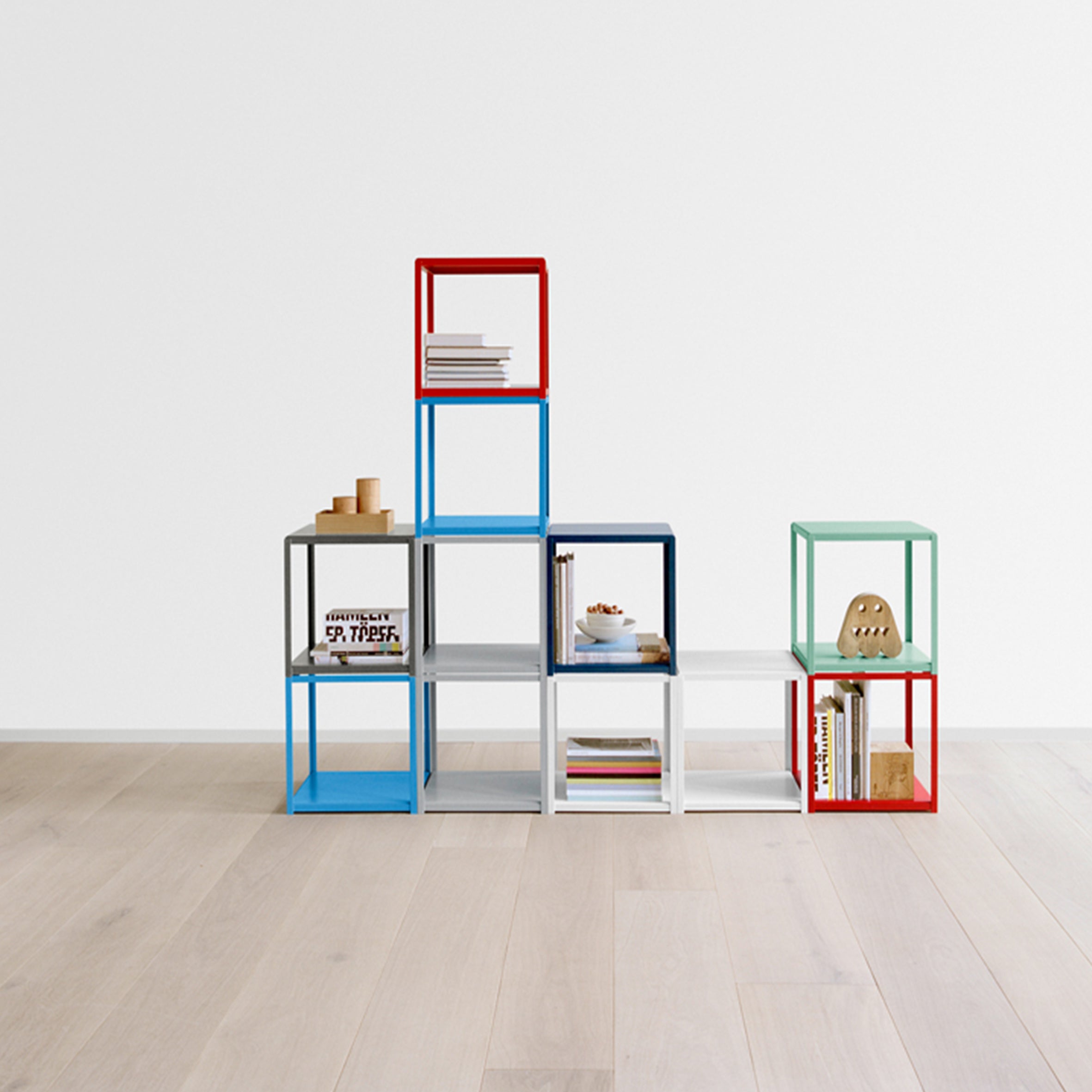 The vibrant FK12 FORTYFORTY shelving unit by E15, featuring open cubes of different heights, showcases books and decor. Made from powder-coated steel, it stands against a white wall on a light wood floor, providing chic and versatile furniture.