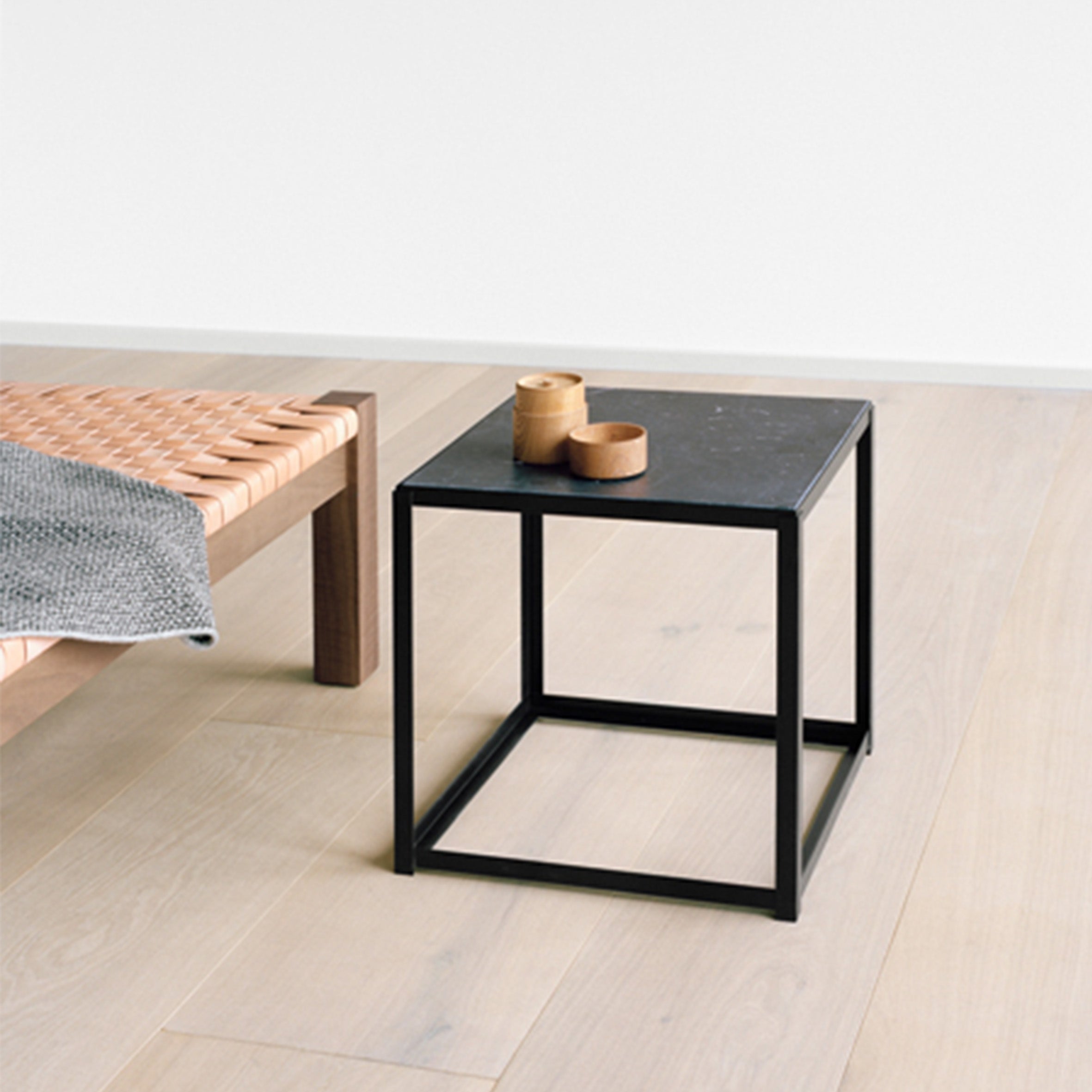 In a minimalist room, an E15 FK12 FORTYFORTY side table with a wooden top holds two small wooden containers. A woven wooden bench with a gray blanket complements the light wood flooring, emphasizing the versatile furniture theme.