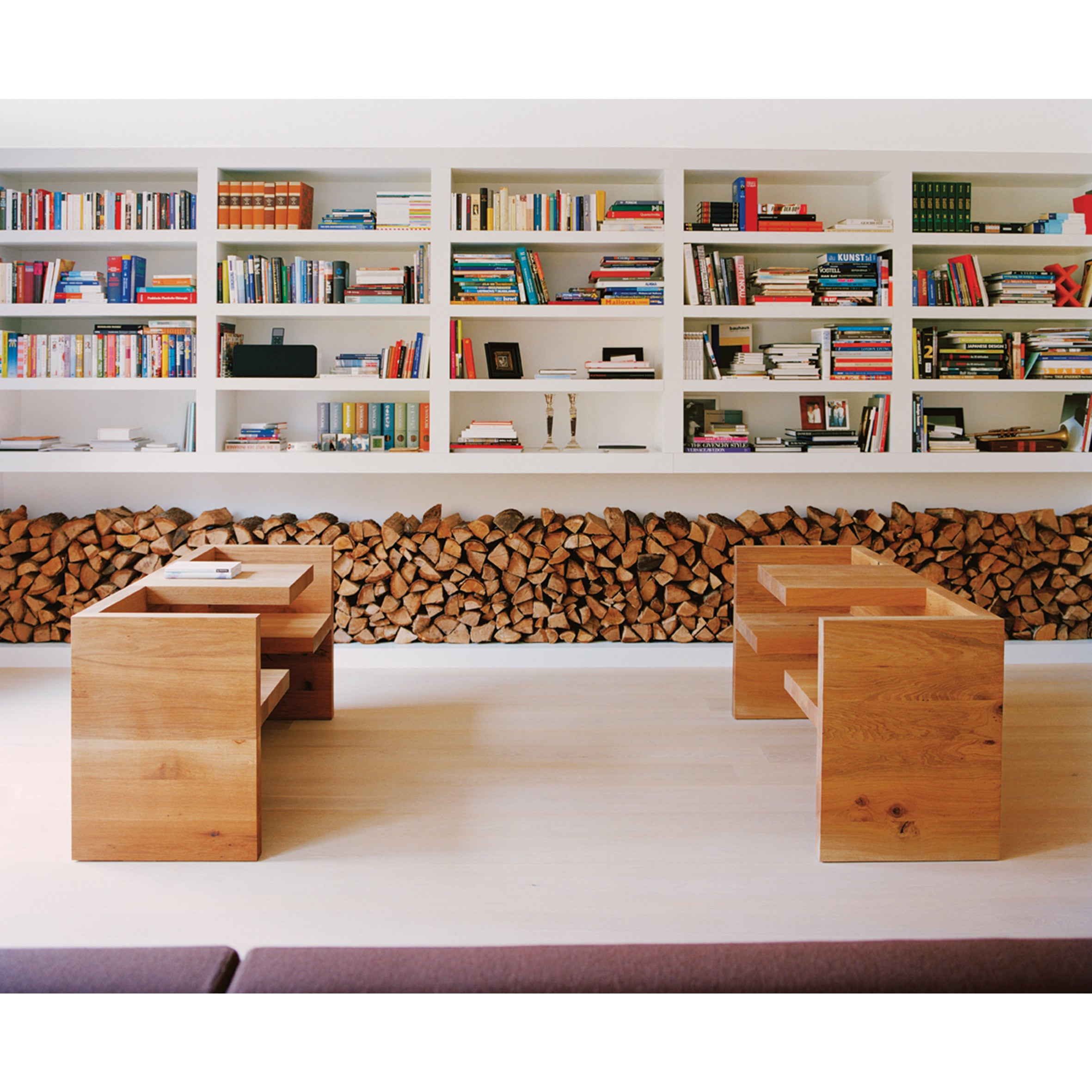 Modern living room with two HP01 TAFEL wooden tables by E15 on a light floor. A wall-length bookshelf filled with books and logs beneath it forms the backdrop, while an aluminum bench enhances the cozy, library-like atmosphere.