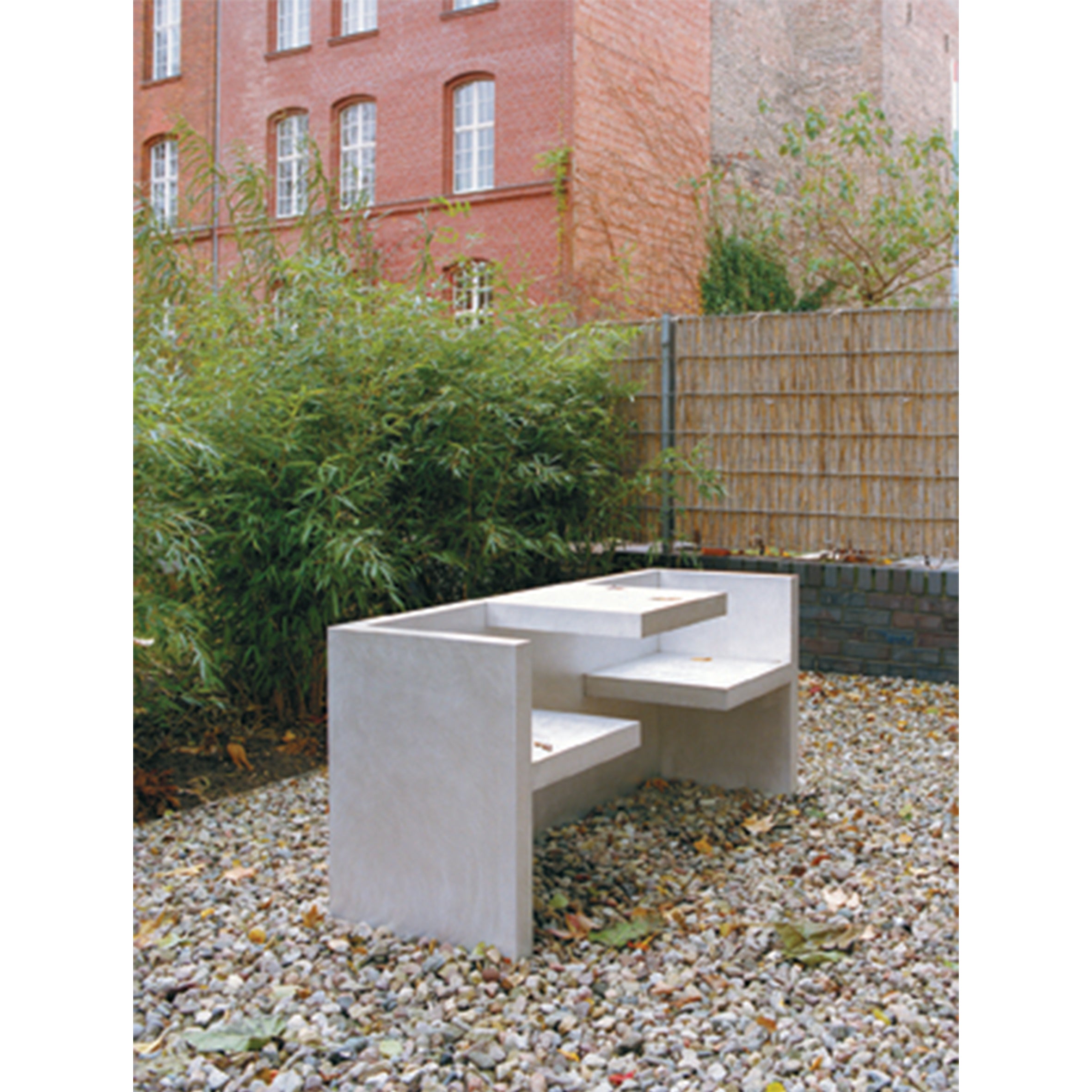 The E15 HP01 TAFEL, a modern concrete table, offers a unique seating experience on a gravel surface outdoors. It is framed by green plants, a brick building with tall windows, and a wooden fence; scattered fallen leaves add to the natural ambiance.