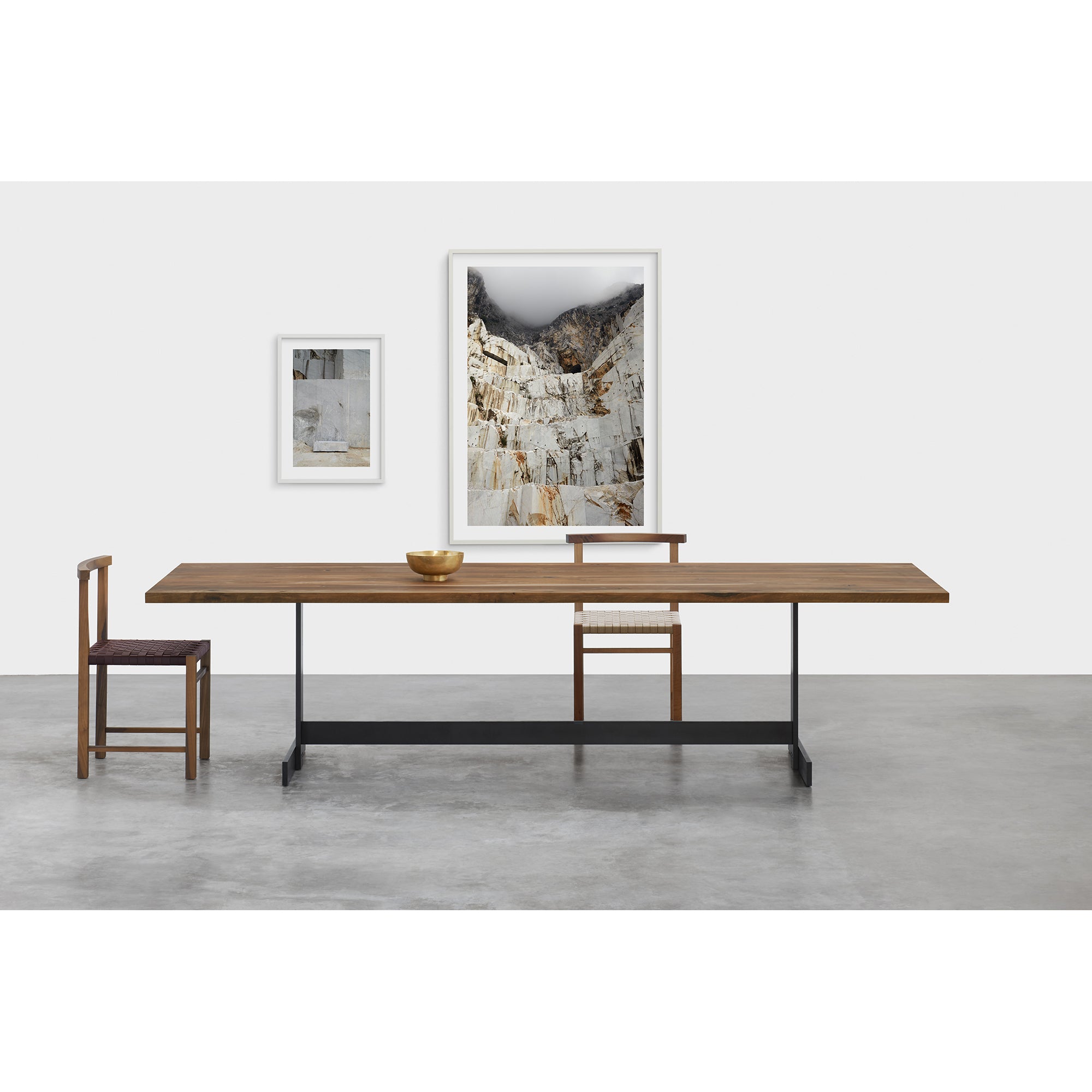 A minimalist dining area features the E15 TA23 KAZIMIR, a long solid European oak table, paired with two chairs. A bowl accents the table, while two framed artworks adorn the white wall—one depicts a rocky landscape. The smooth gray concrete floor complements the rooms clean lines and geometric forms.