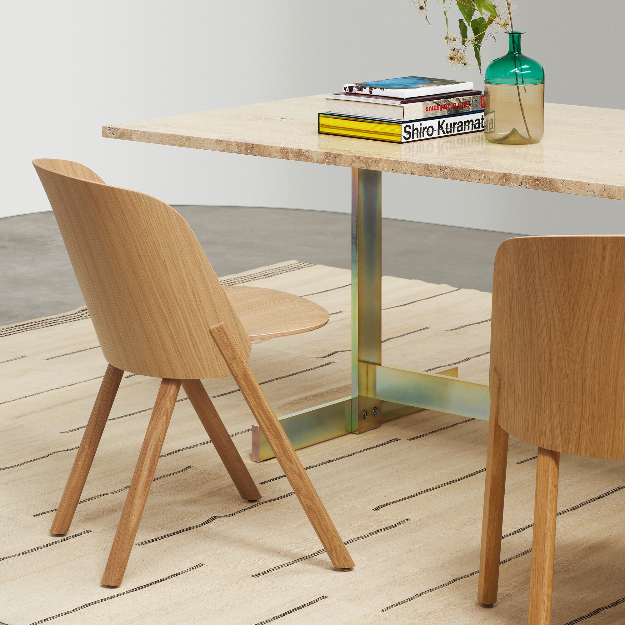 A minimalist dining area showcases the E15 TA23 KAZIMIR table made of solid European oak on a striped rug. Two wooden chairs with curved backs flank the table, which holds a green vase, books, and a small plant.