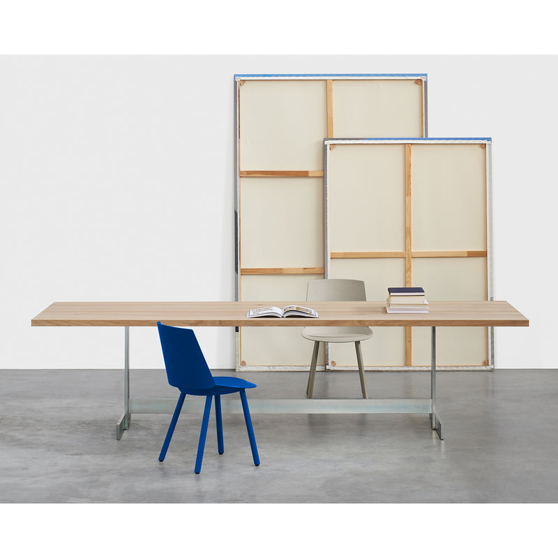 A minimalist room showcases the E15 TA23 KAZIMIR table made of solid European oak, flanked by a blue chair at the front and a beige one in the back. Books and an open magazine lie on the table, while two large blank canvases bring geometric shapes to the white wall.