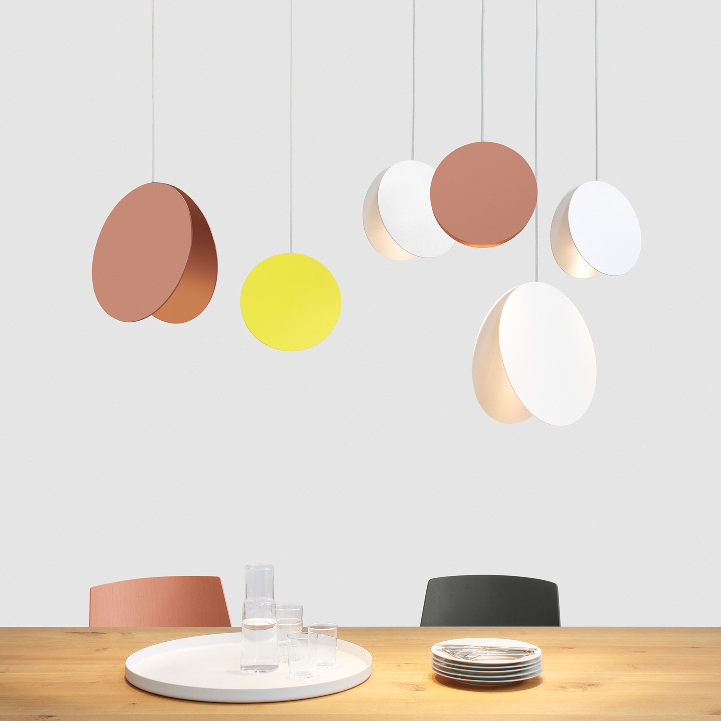 A modern dining area features a wooden table with an elegant white tray, glassware, and plates. Overhead, E15s LT05 NORTH Pendant Light - Sale in brown, yellow, and white casts a poetic light like lunar phases. Two chairs are partially visible in the background.