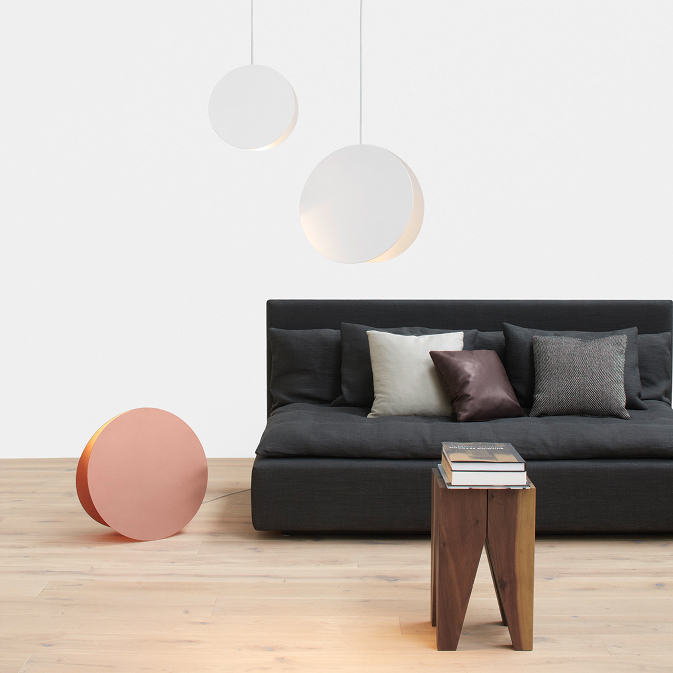 A minimalist living room showcases a dark gray sofa with gray and beige pillows. Three pendant lights provide atmospheric lighting, complemented by the E15 LT05 NORTH Floor Light on the floor. A wooden side table with books rests on a light wooden floor echoing abstract geometry.