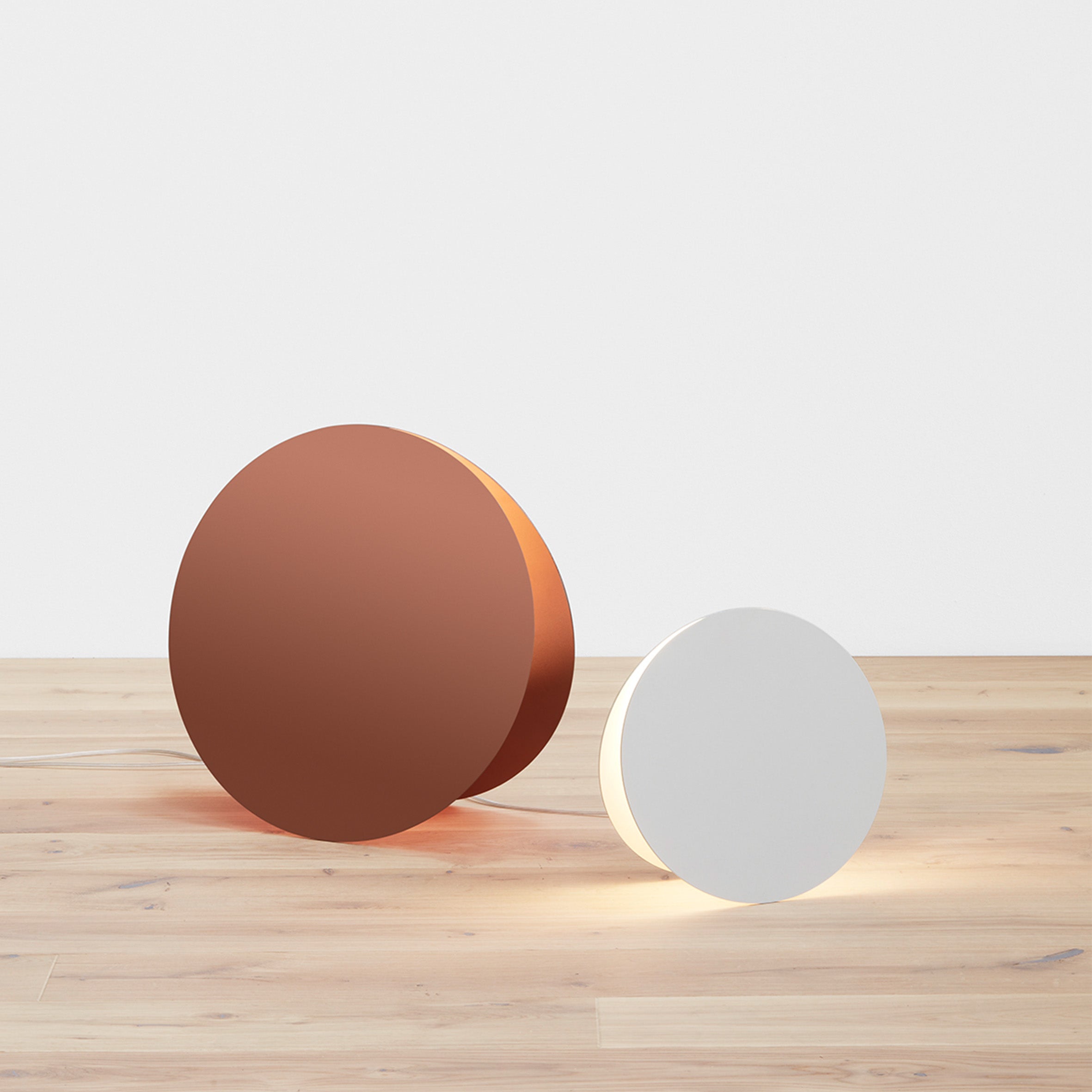 The LT05 NORTH Floor Light by E15 is featured in a minimalist setting with abstract geometry. Displayed are two round objects on a wooden floor against a white backdrop, a larger copper-colored lamp and a smaller white one, both emitting soft, sculptural light.