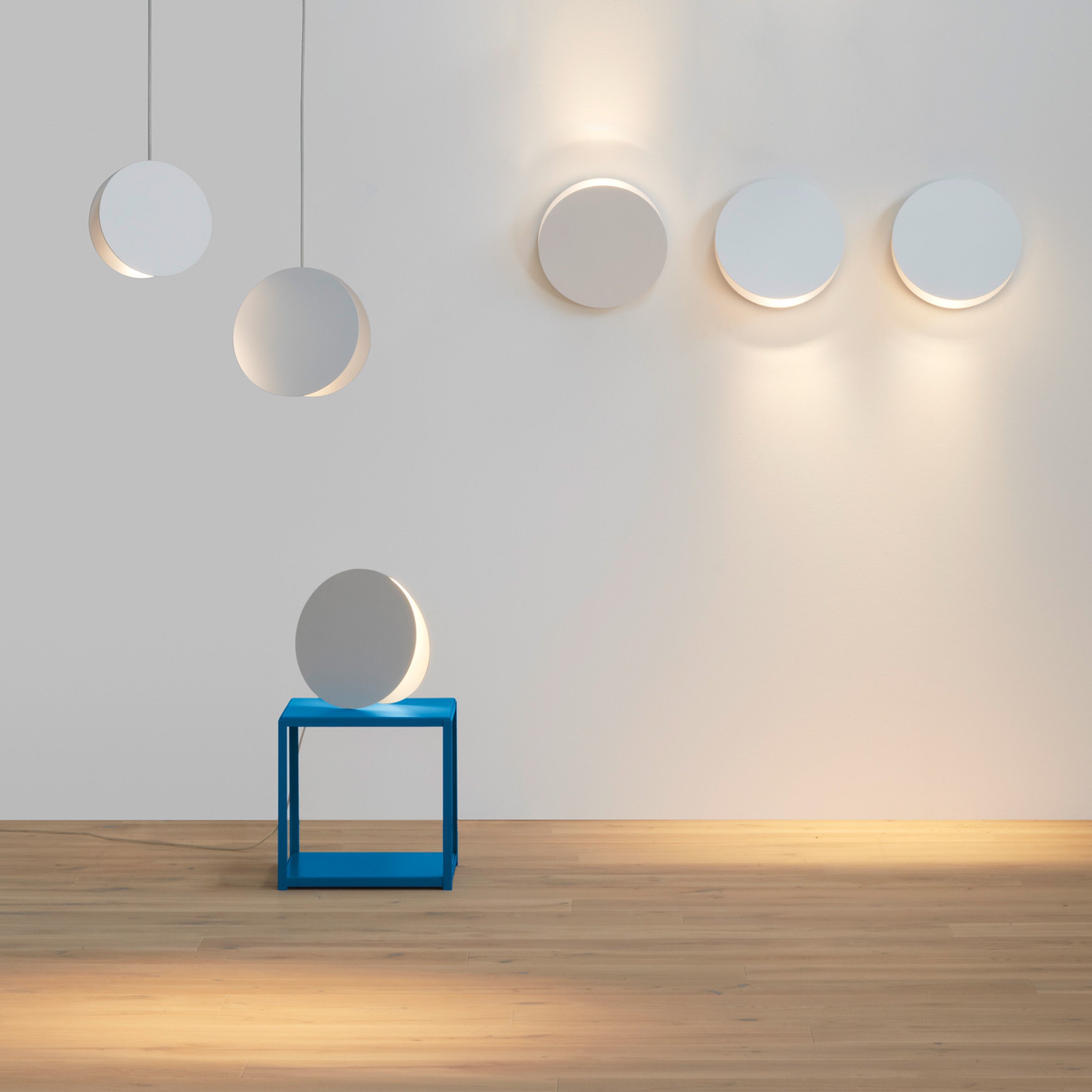 A minimalist room features circular wall lights and two pendant lights on a white wall. The E15 LT05 NORTH Floor Light Sale edition sits sculpturally on a small blue table, adding character on the light wood floor. Atmospheric lighting casts soft shadows and warm tones throughout the space.