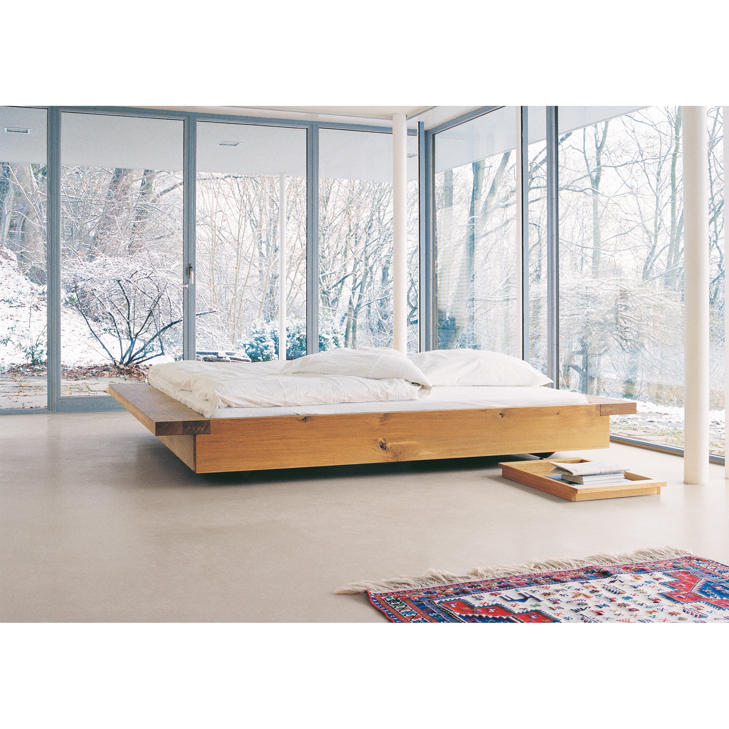 A minimalist bedroom showcases the E15 PA02 NOAH solid wood platform bed with white bedding. Large windows offer views of a snowy forest. A colorful rug accents the light floor near the bed, and a small wooden tray holding a book completes this serene retreat amidst nature.