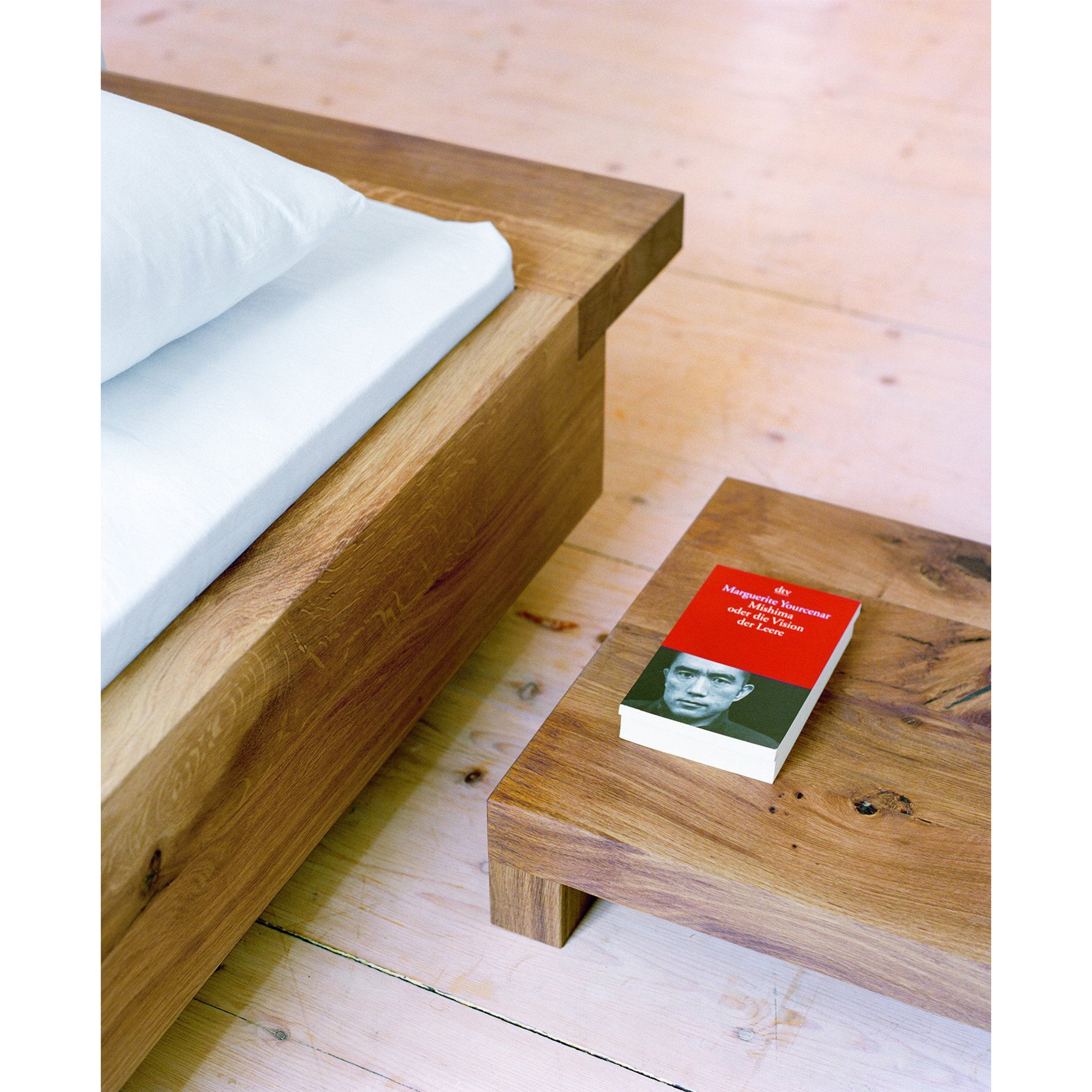 A minimalist room features E15s PA02 NOAH solid wood bed with white bedding and a matching wooden side table. On the table, theres a book with a red cover and black-and-white portrait. The light wood floor complements the beds adjustable feet for perfect balance.