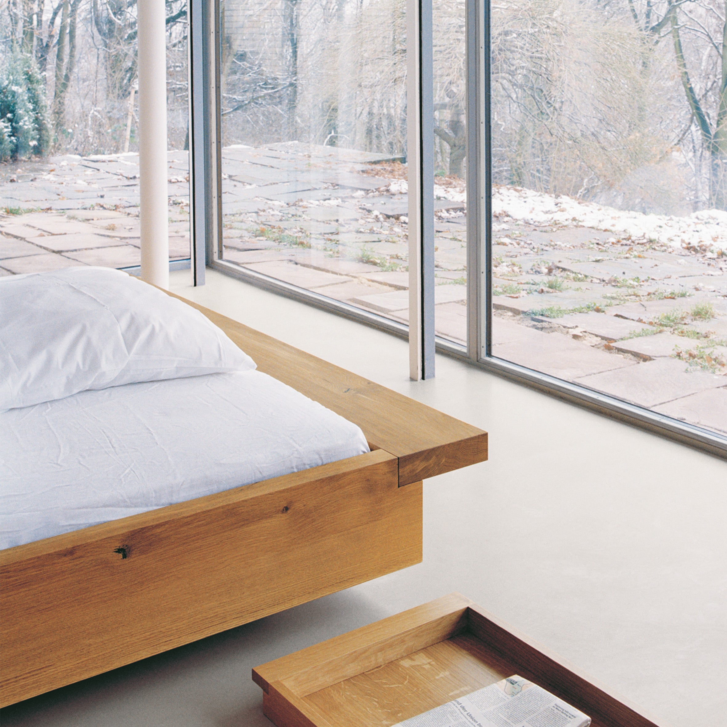 A minimalist bedroom includes the low PA02 NOAH solid wood bed frame by E15 with white bedding. Floor-to-ceiling glass windows open to a patio, filling the bright room with natural light, while a wooden tray sits on the floor next to the bed.
