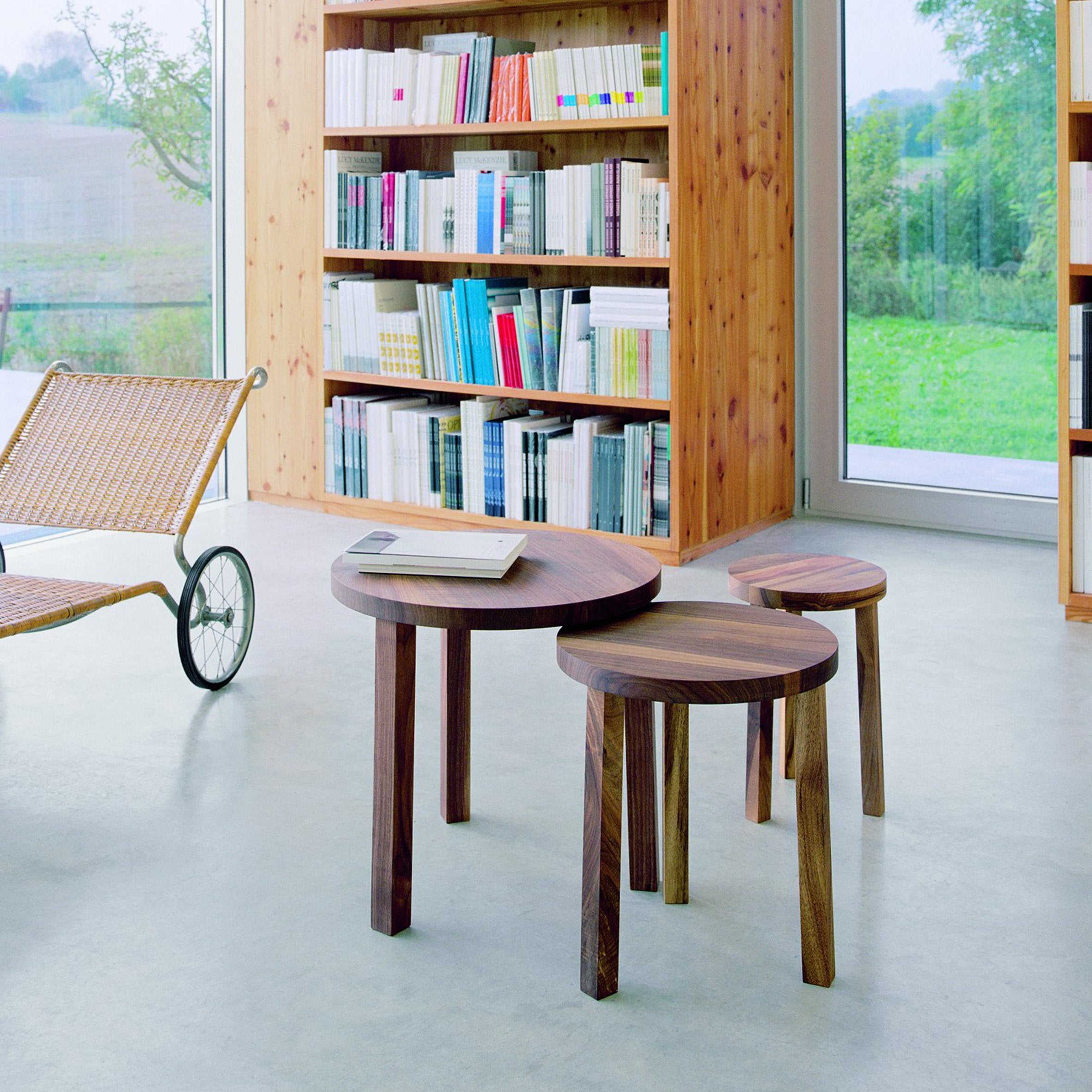 A cozy room features a set of E15 PA03 ALEX tables with solid wood tops, a wheeled wicker lounge chair, and a large bookshelf filled with colorful books. The expansive window offers views of the verdant landscape outside.