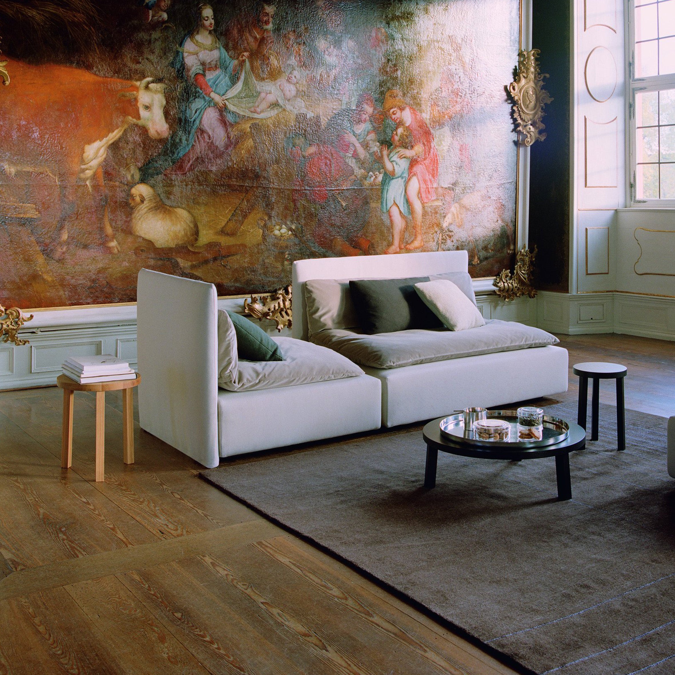 A stylish room showcases a large mural of historical figures. A modern white sofa with gray cushions pairs with the PA05 LEILA coffee table by E15, crafted from solid European oak, and a wooden side table on a brown rug. Natural light filters in.