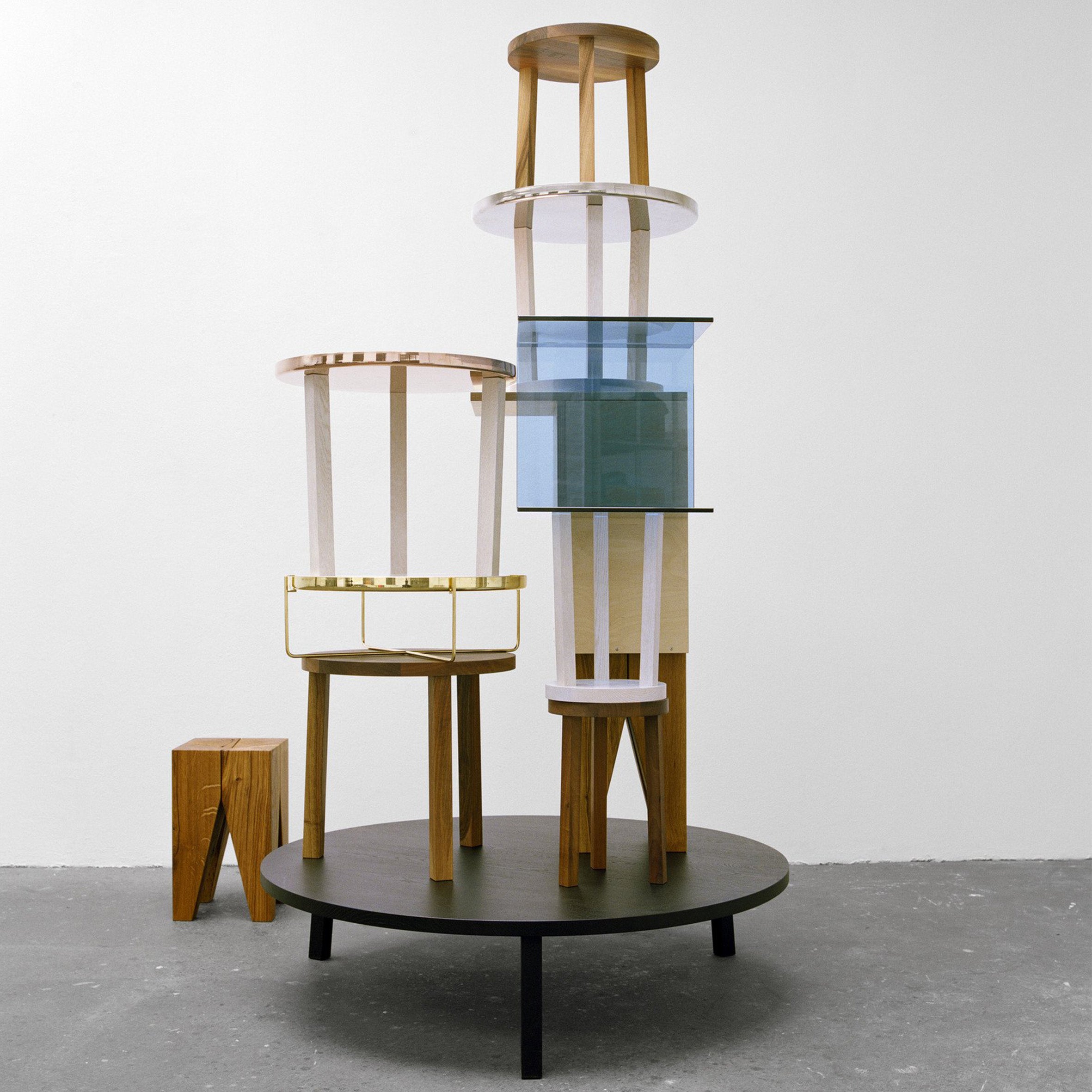 The modern art sculpture consists of stacked circular wooden tables and stools in solid European oak, complemented by translucent white and blue glass panels. It stands on a large black platform in a minimalist white space, reminiscent of E15s PA05 LEILA coffee table designs.