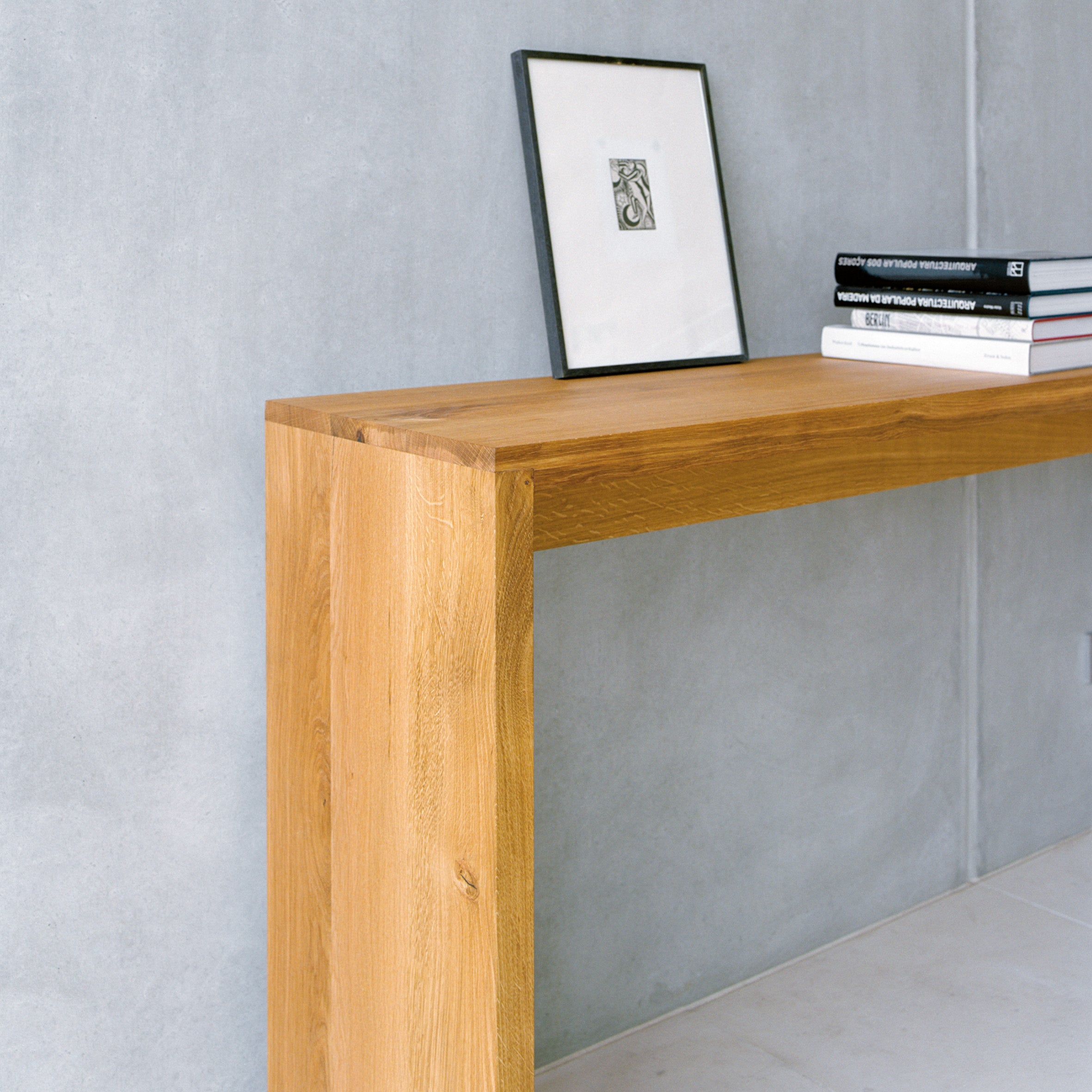 The minimalist wooden console table against a gray wall showcases the elegance of E15 design by Philipp Mainzer. A framed picture leans on the wall, complemented by three stacked books, enhancing the modern and clean aesthetic inspired by the SB01 ALTO sideboard.