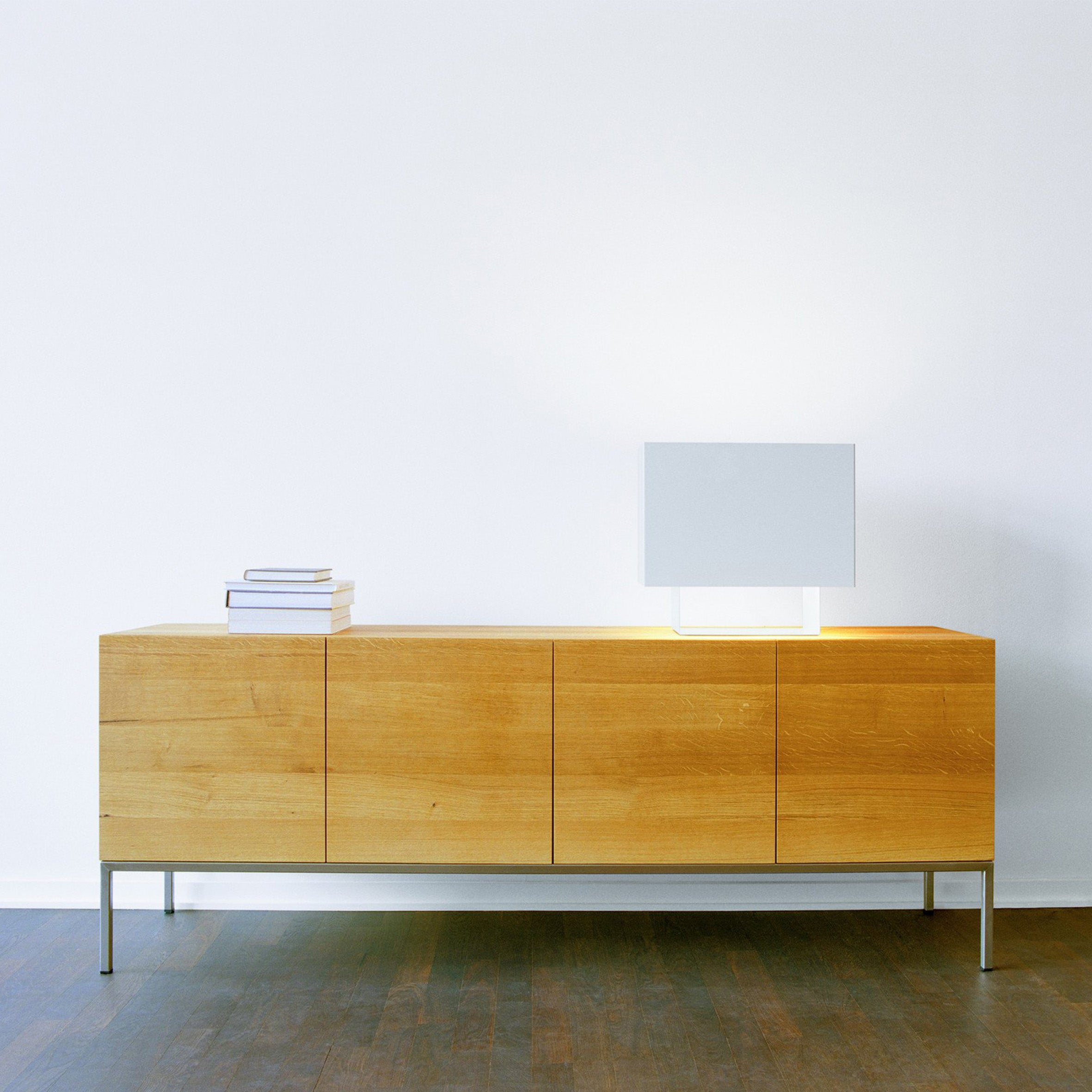 A minimalist room showcases the E15 SB02 FARAH, a solid wood sideboard with metal legs, against a white wall. On it rests a modern square lamp and a small stack of books. The dark wood finish floor is detailed with aluminum strips.