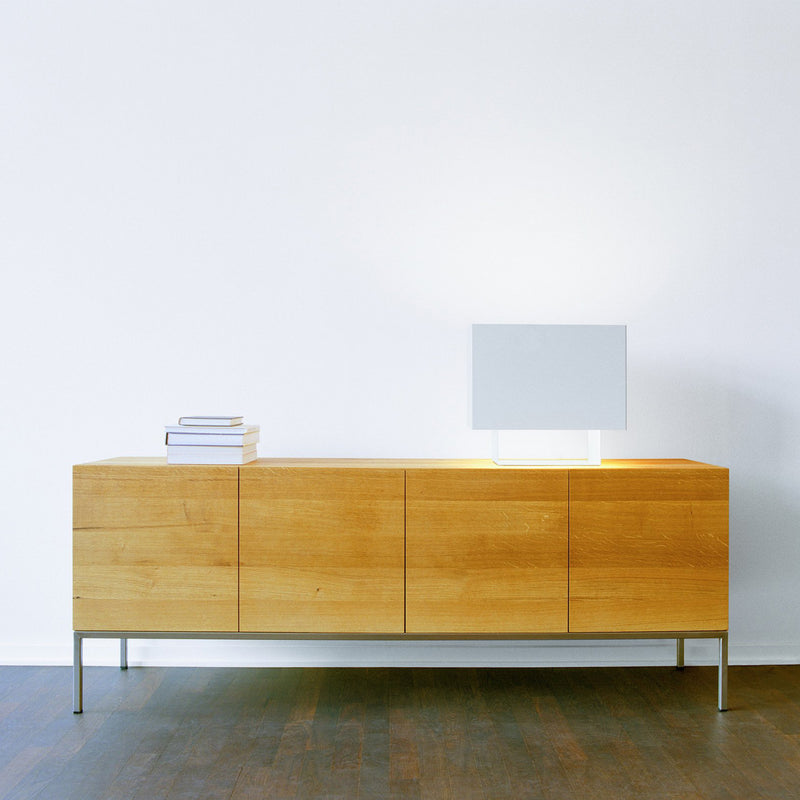 A minimalist room showcases the E15 SB02 FARAH, a solid wood sideboard with metal legs, against a white wall. On it rests a modern square lamp and a small stack of books. The dark wood finish floor is detailed with aluminum strips.