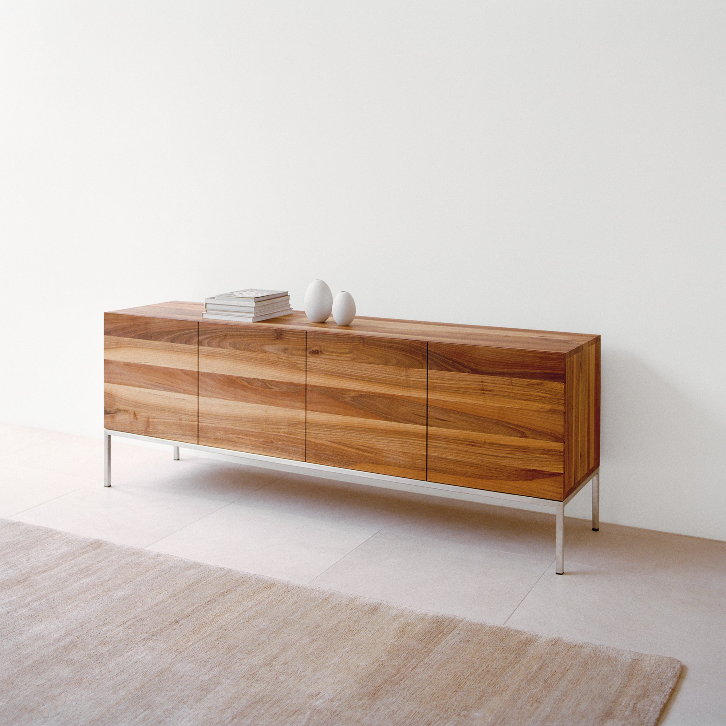 The SB02 FARAH by E15, a minimalist solid wood sideboard with brushed stainless steel legs, stands against a light wall. Clean lines complement two white vases and books on top. A beige rug contrasts beautifully with the light-colored tiles below.