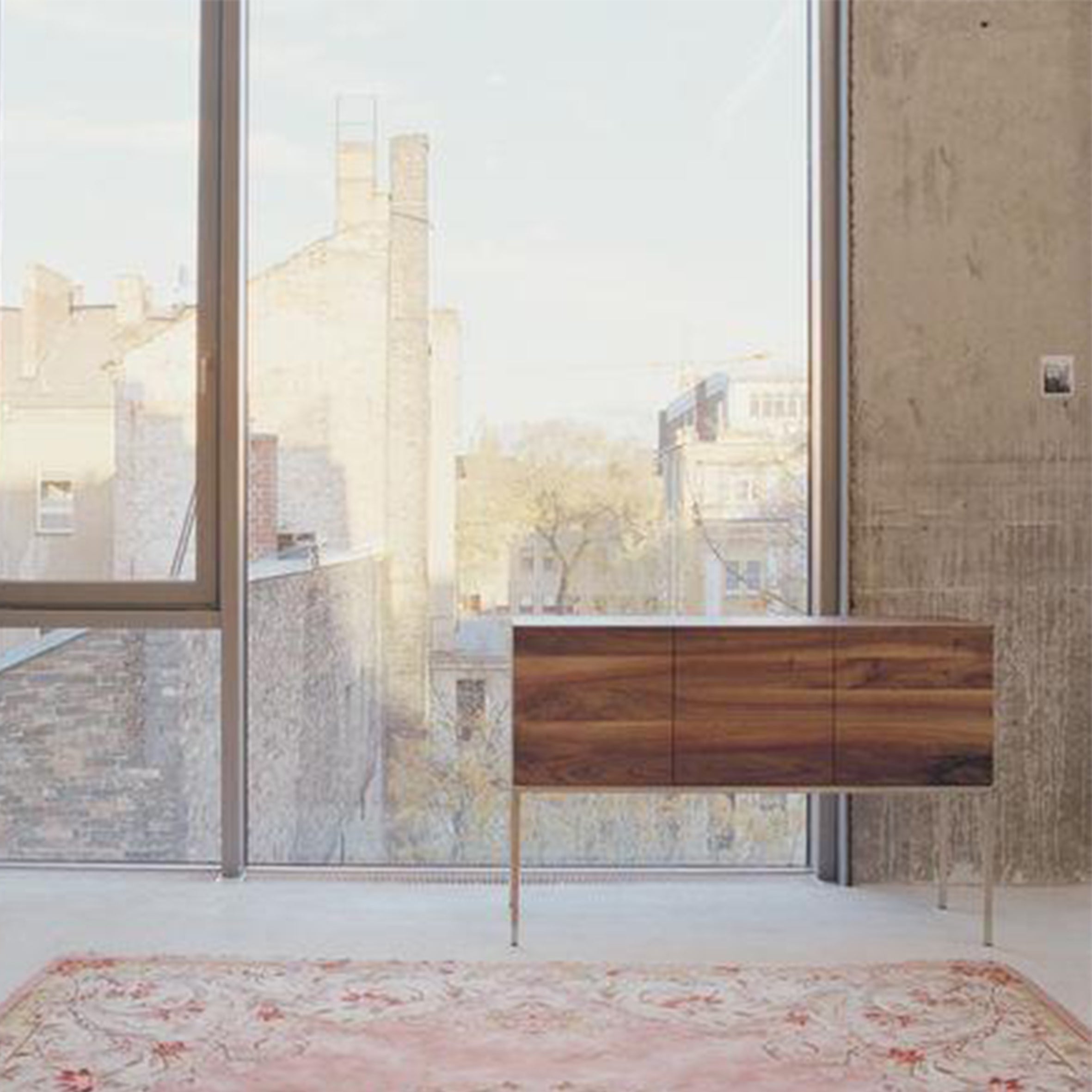 A contemporary room with a large floor-to-ceiling window showcasing a cityscape. The E15 SB02 FARAH solid wood sideboard rests on the light-colored floor, complemented by a pastel decorative rug. An unfinished concrete wall beside the window adds an industrial touch.