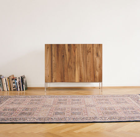 A minimalist room showcases the E15 SB03 FARIBA solid wood cabinet with automatic closure against a plain white wall, a stack of books on the floor to the left, and a patterned rug partially covering the wooden parquet floor.