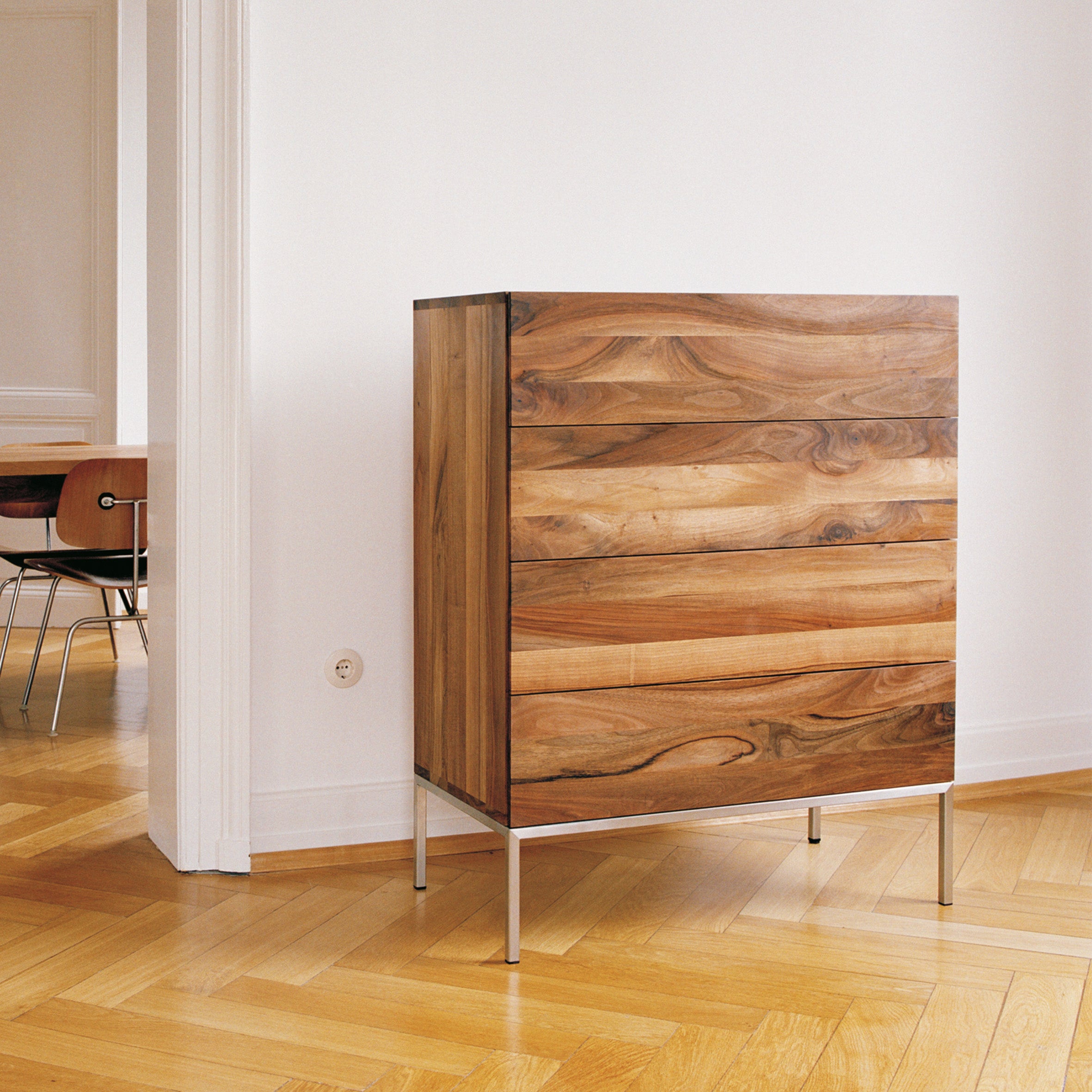 The SB04 FATIMA dresser by E15, featuring a natural varnished finish and four drawers, stands on brushed stainless steel legs against a white wall. It rests on a light brown herringbone wood floor, with part of a solid wood chair at an adjoining table visible.