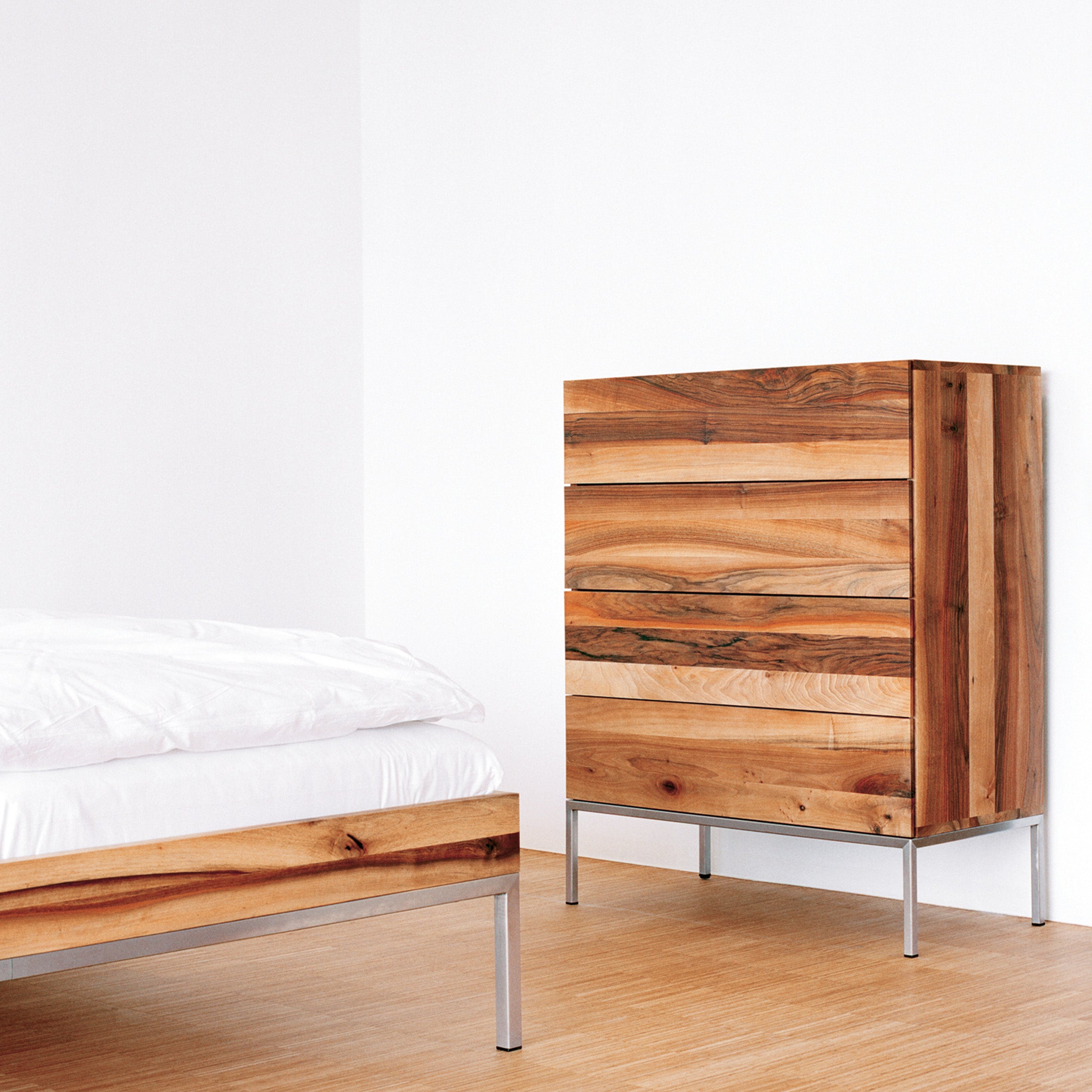 A minimalist bedroom showcases the E15 SB04 FATIMA solid wood dresser and bed, both with brushed stainless steel legs. The bed is styled with a white mattress and sheets, complemented by a light wood floor and plain white walls for a sleek, modern aesthetic.