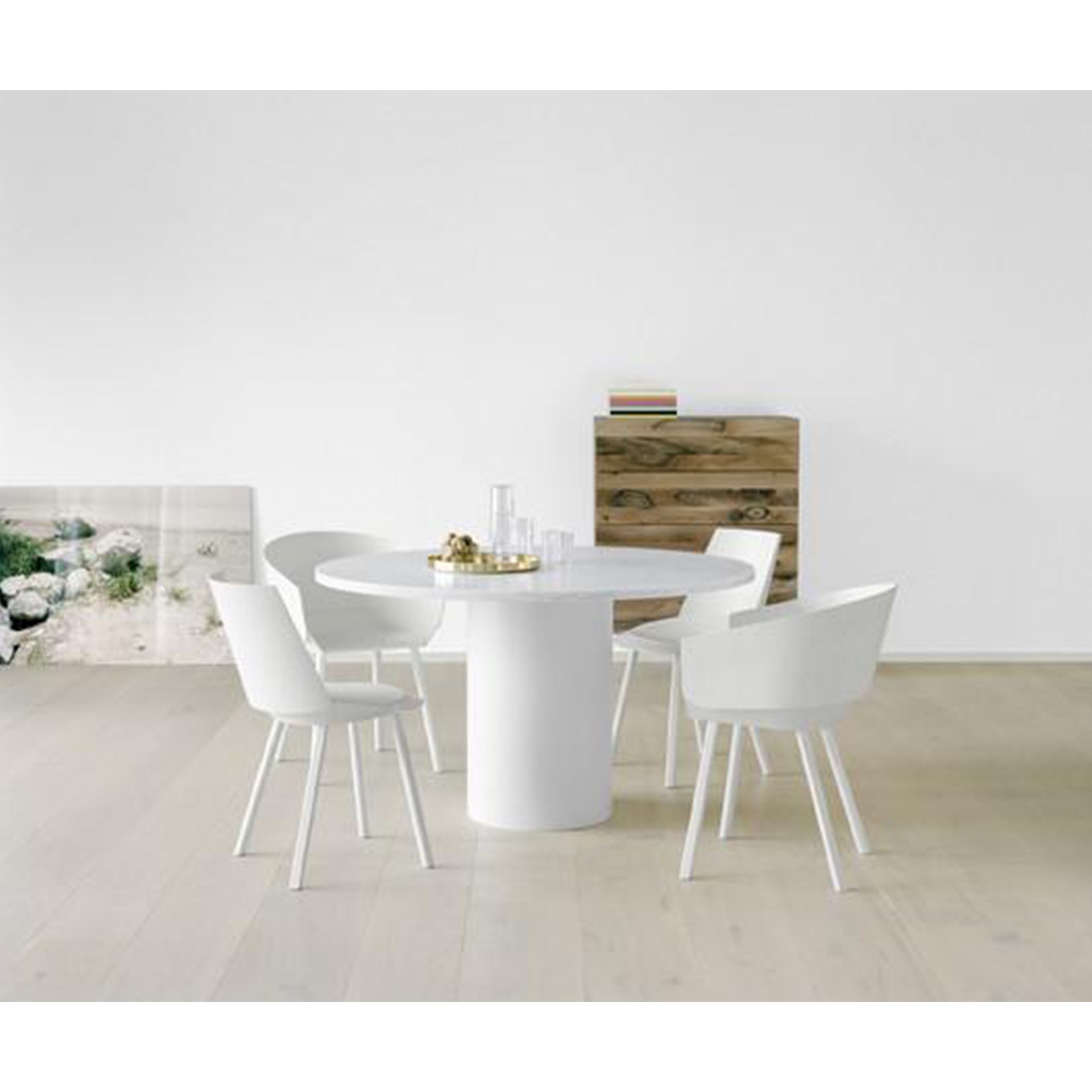 Minimalist dining room featuring the SB04 FATIMA round white table by E15, with adjustable feet and four white chairs on light wood flooring. A solid wood cabinet, accompanied by stacked books and a large artwork leaning against the wall, completes the serene setting.