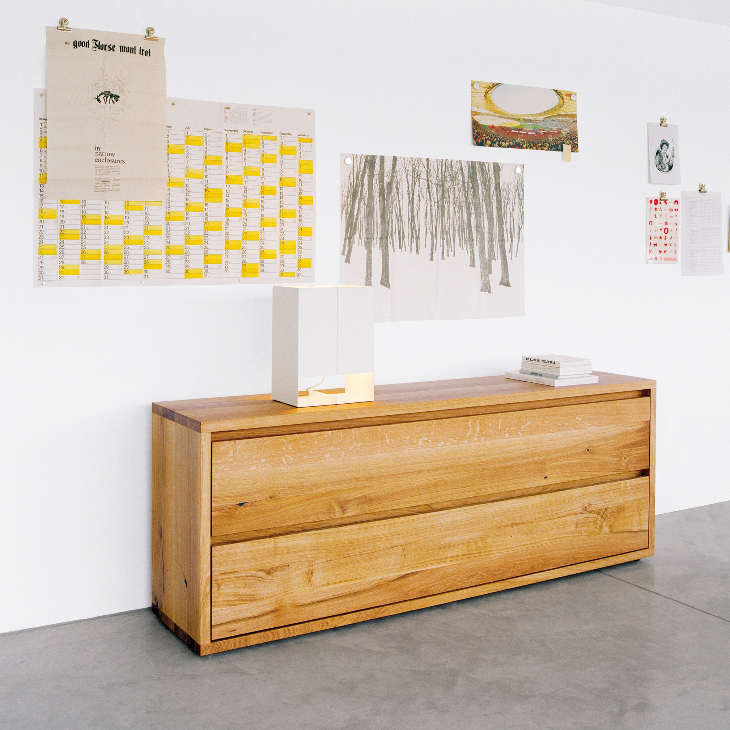 A minimalist room features an E15 SB05 IMARI oak sideboard with two drawers against a white wall, adorned with clipped paper artworks and a calendar. A modern lamp and book are elegantly arranged on its solid wood surface, while the floor boasts polished concrete.