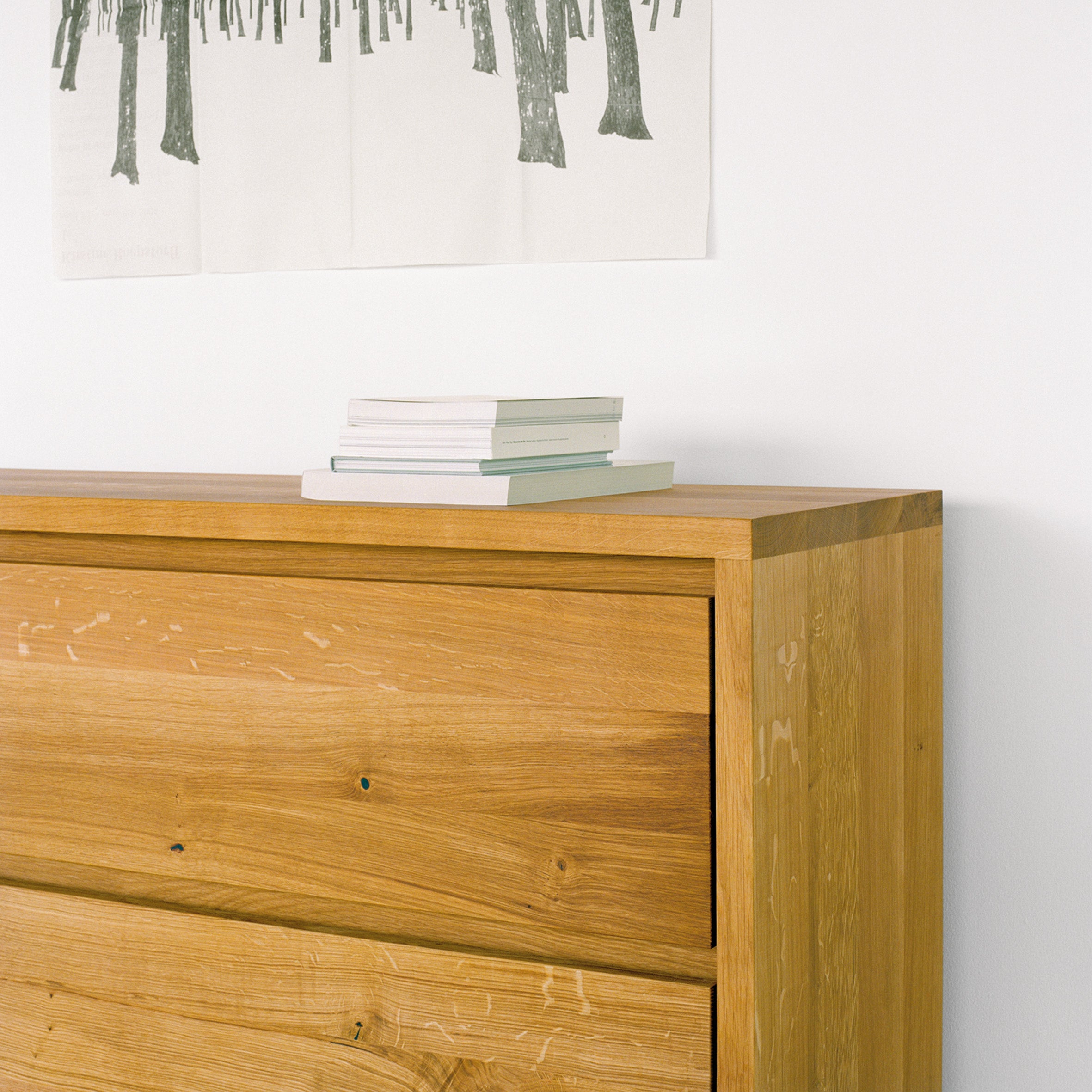 An E15 SB05 IMARI solid oak chest of drawers sits against a white wall, topped with neatly stacked books. Above it hangs a wall poster featuring a green tree design, perfectly complementing the elegant furniture below.